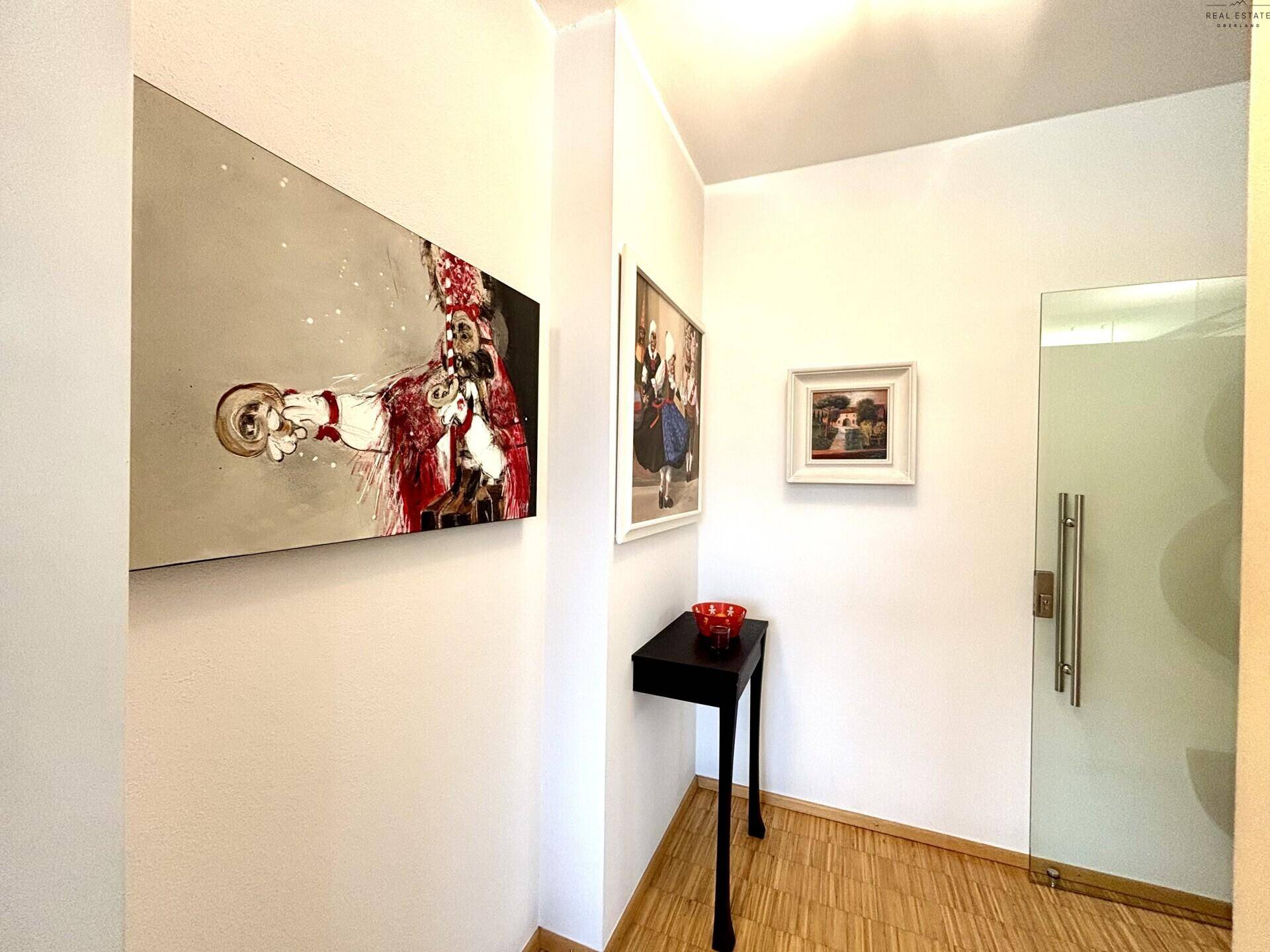 Heller Flur mit Kunstwerken, einem kleinen Beistelltisch und einer Glastür.