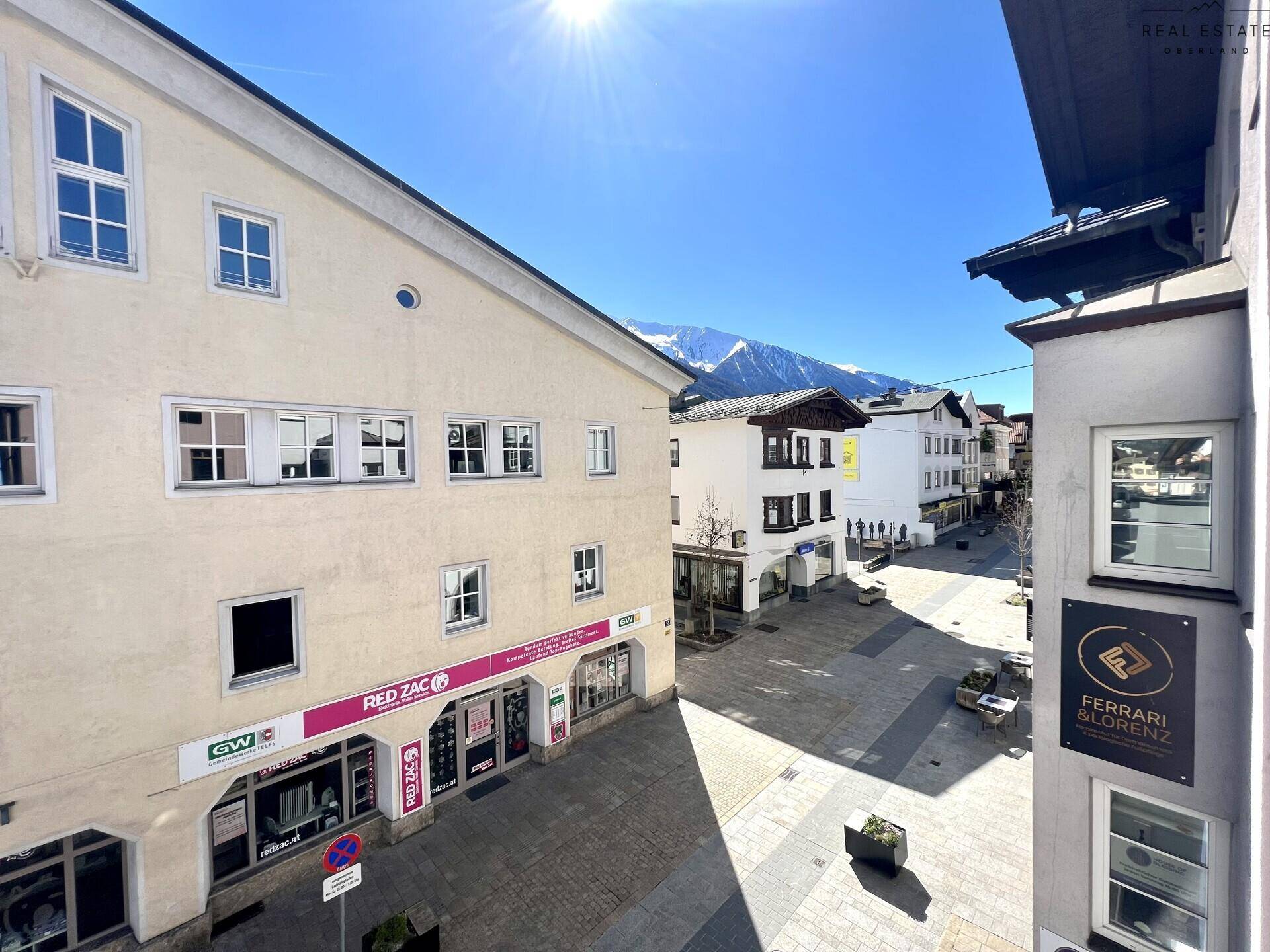 Sonniger Blick auf die Einkaufsstraße mit Geschäften und den majestätischen Bergen im Hintergrund.