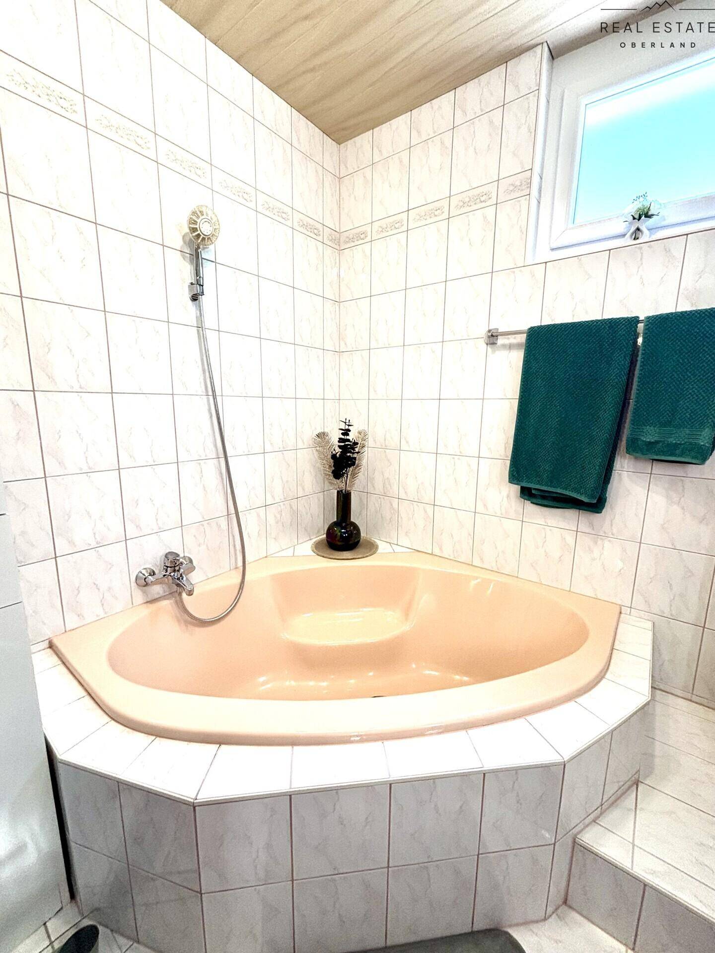 Badezimmer mit Eckbadewanne und Duschkopf, hellen Fliesen und einem Fenster für Tageslicht.