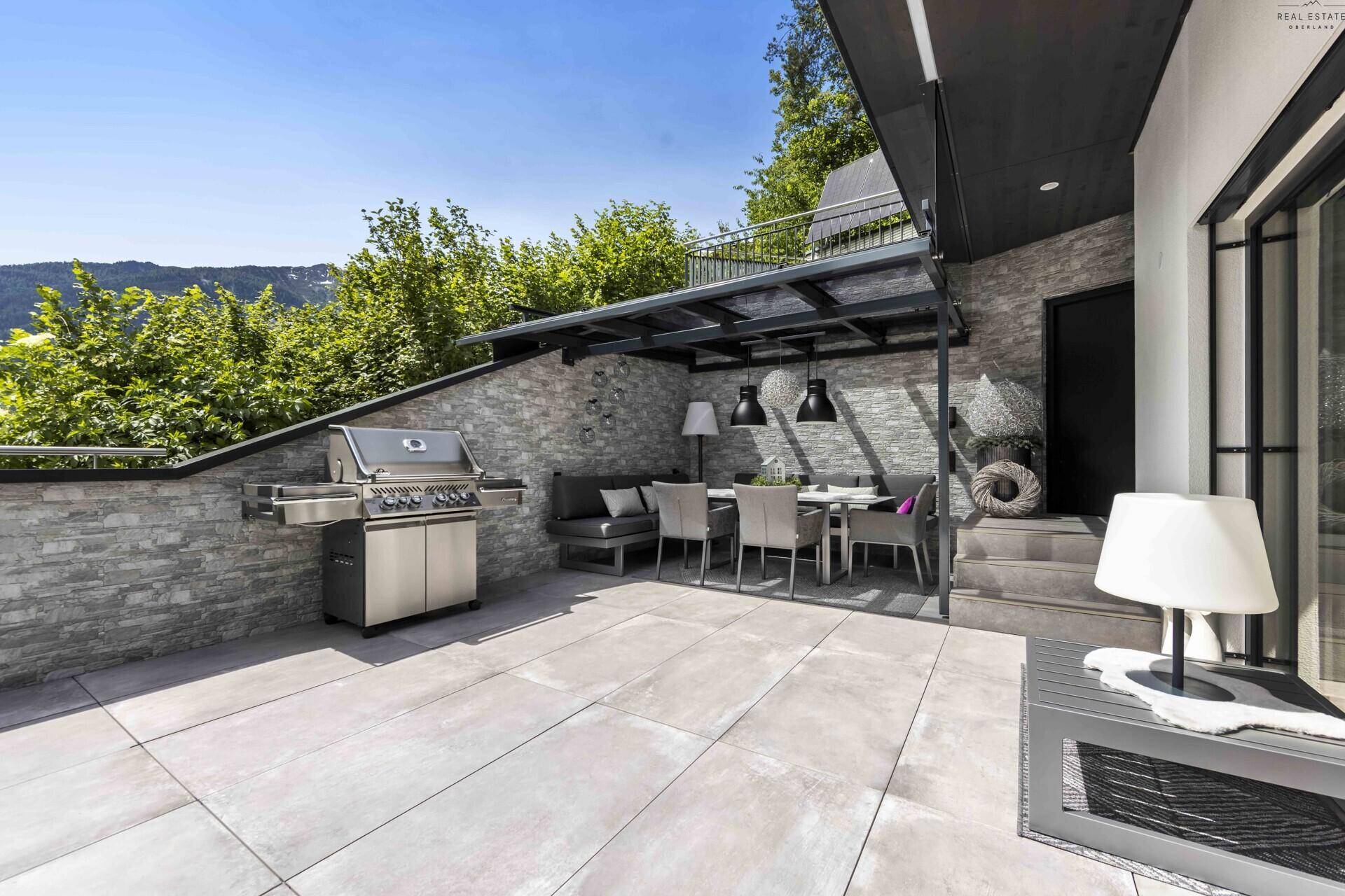 Moderne Terrasse mit Grillbereich und Esstisch, umgeben von grüner Natur und Bergblick.