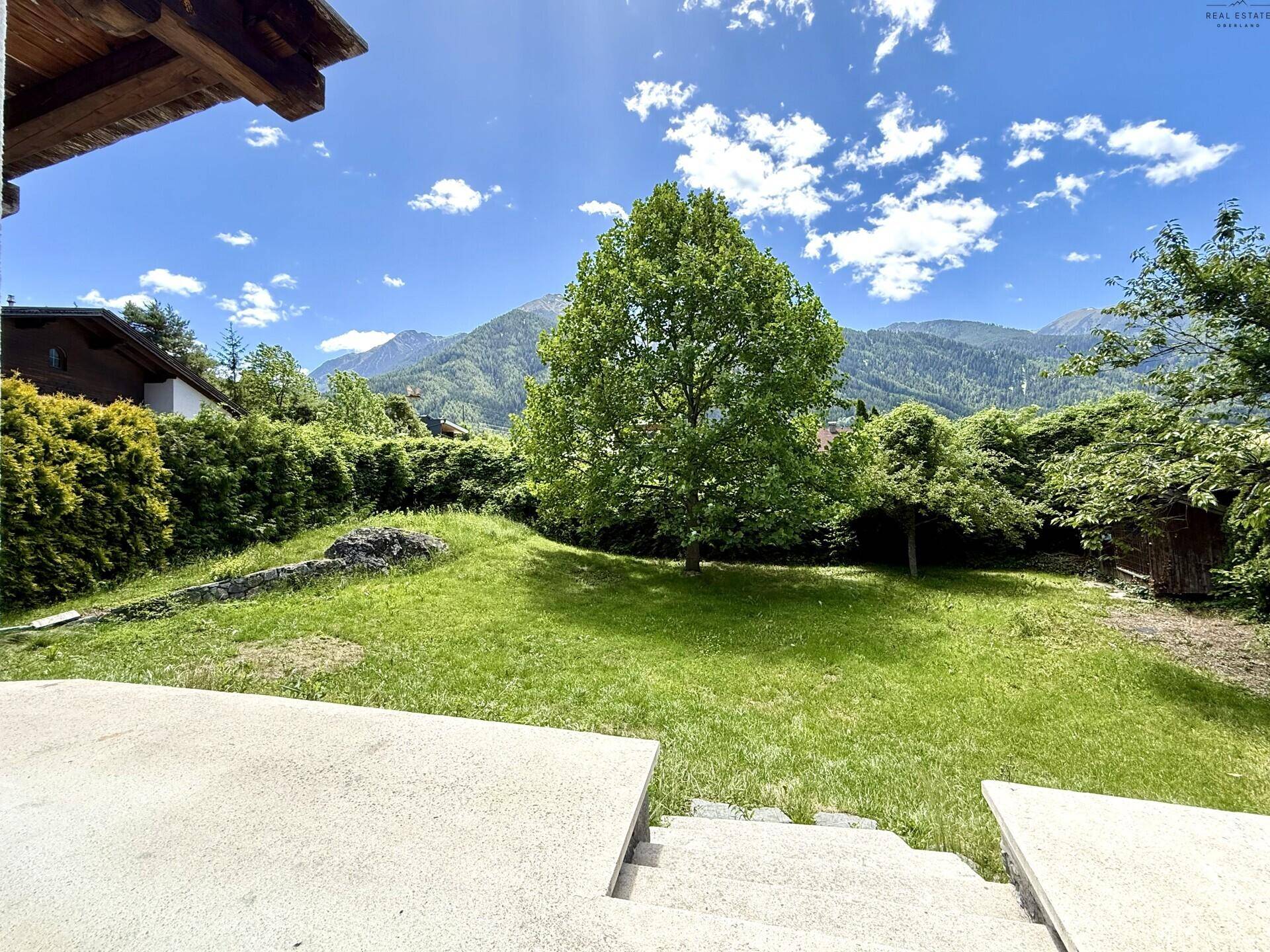 Sonniger Garten mit Rasenfläche, großem Baum und Blick auf die umliegenden Berge.