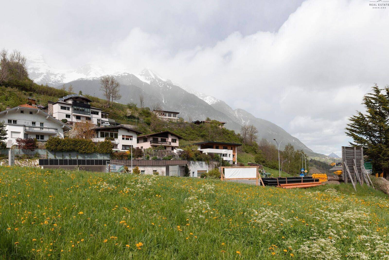 Eine grüne Wiese mit Häusern am Hang und schneebedeckten Bergen unter bewölktem Himmel.