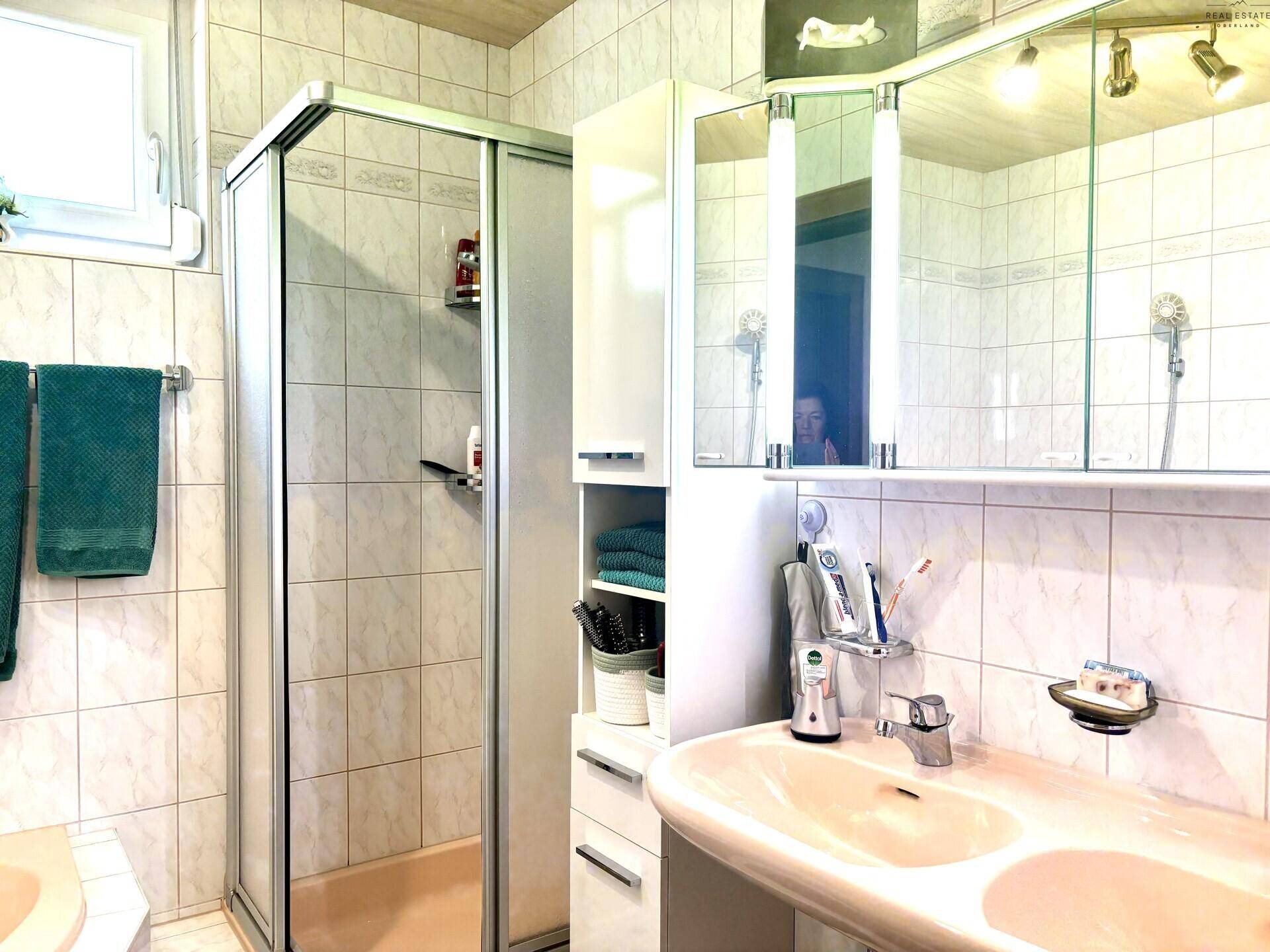 Badezimmer mit Duschkabine, Einzelwaschbecken und Spiegelschrank, hell gefliest.