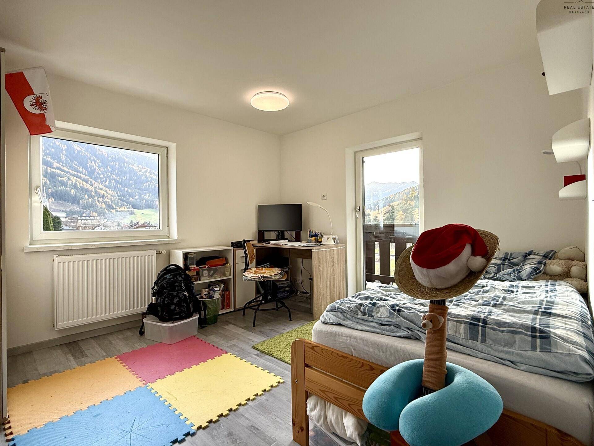 Kinderzimmer mit Bett, Schreibtisch, bunten Spielmatten und Balkonzugang.