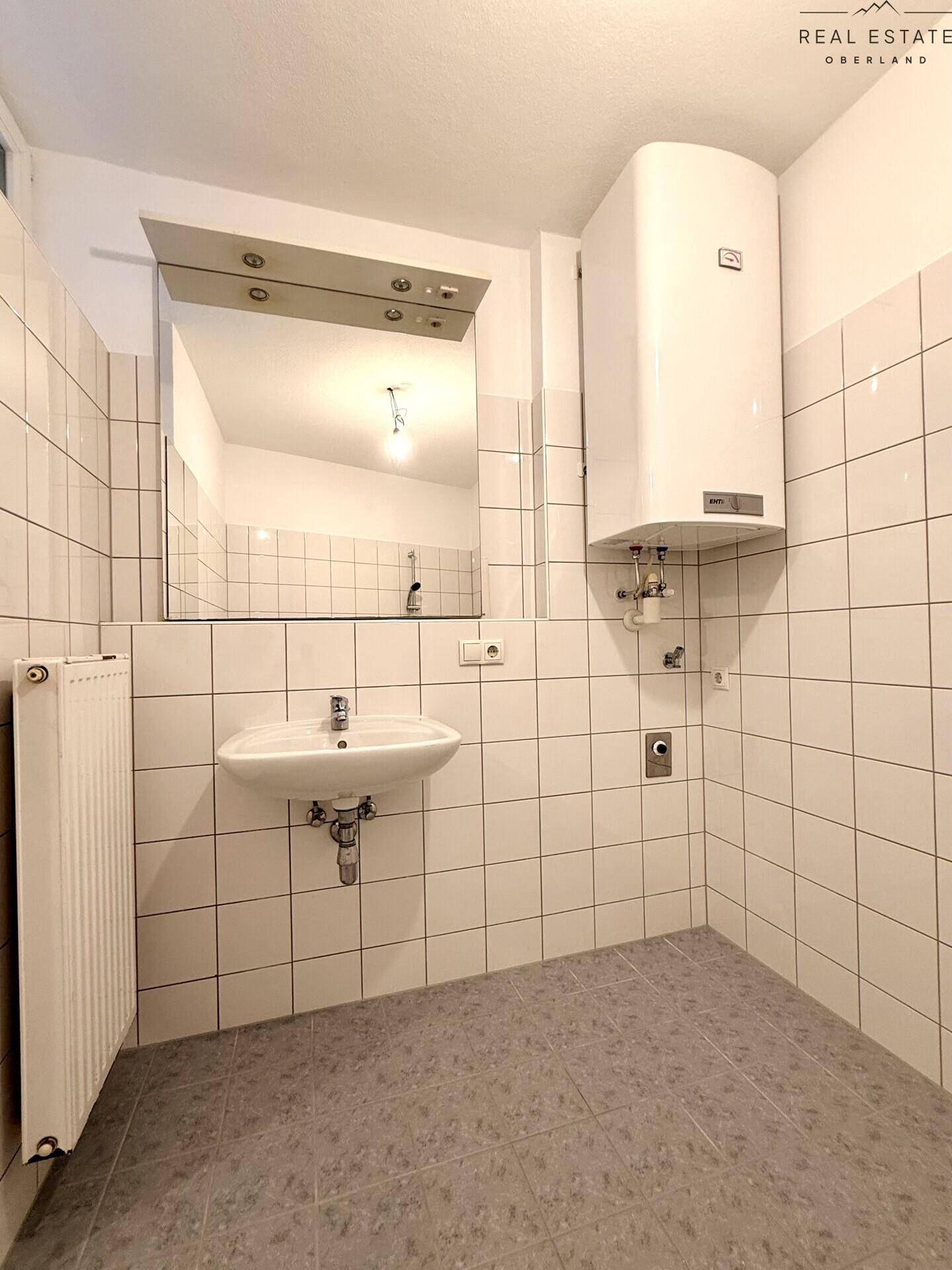 Badezimmer mit weißen Fliesen, Waschbecken und Warmwasserboiler.