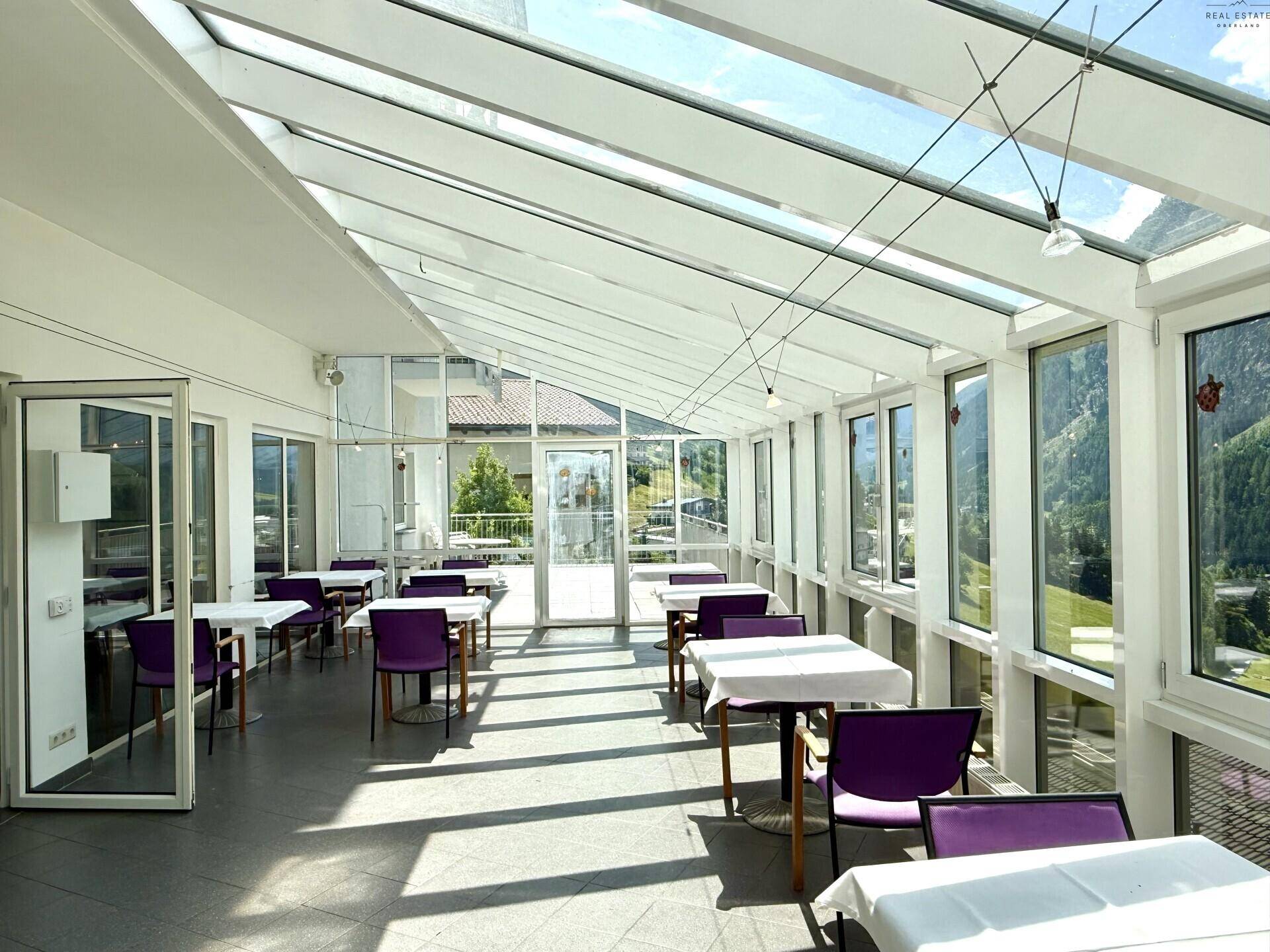 Lichtdurchfluteter Wintergarten mit vielen Tischen und Stühlen, ideal für Mahlzeiten mit Bergblick.