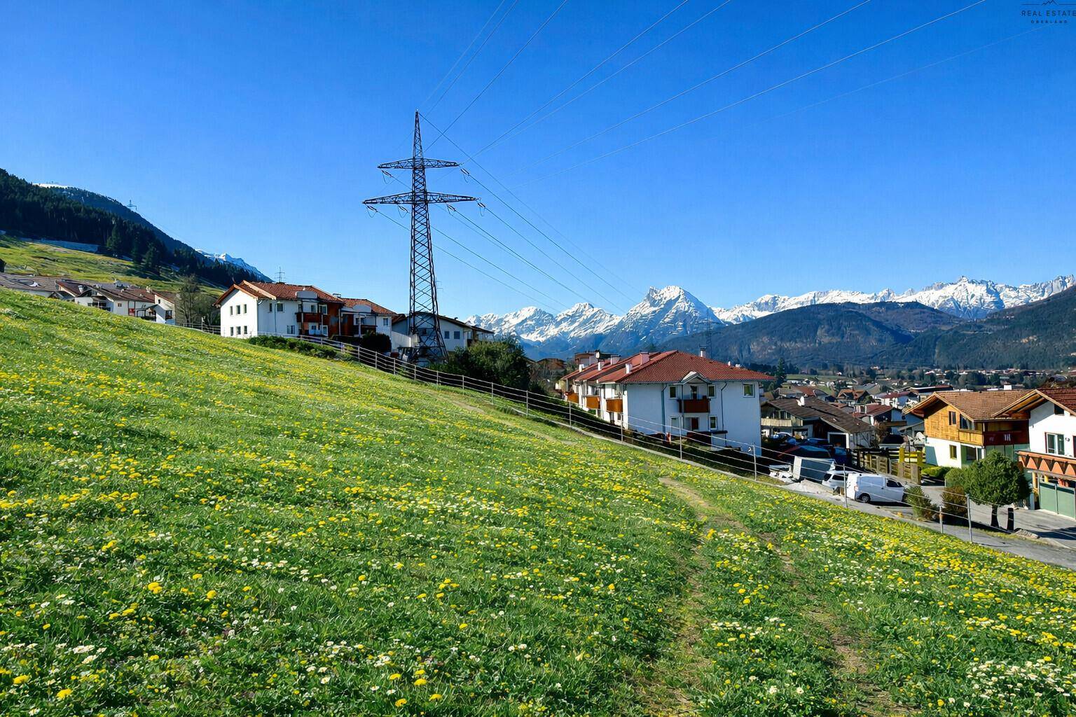 Hügelige Landschaft mit blühenden Wiesen, Wohngebäuden und einem weiten Blick auf die Alpen.