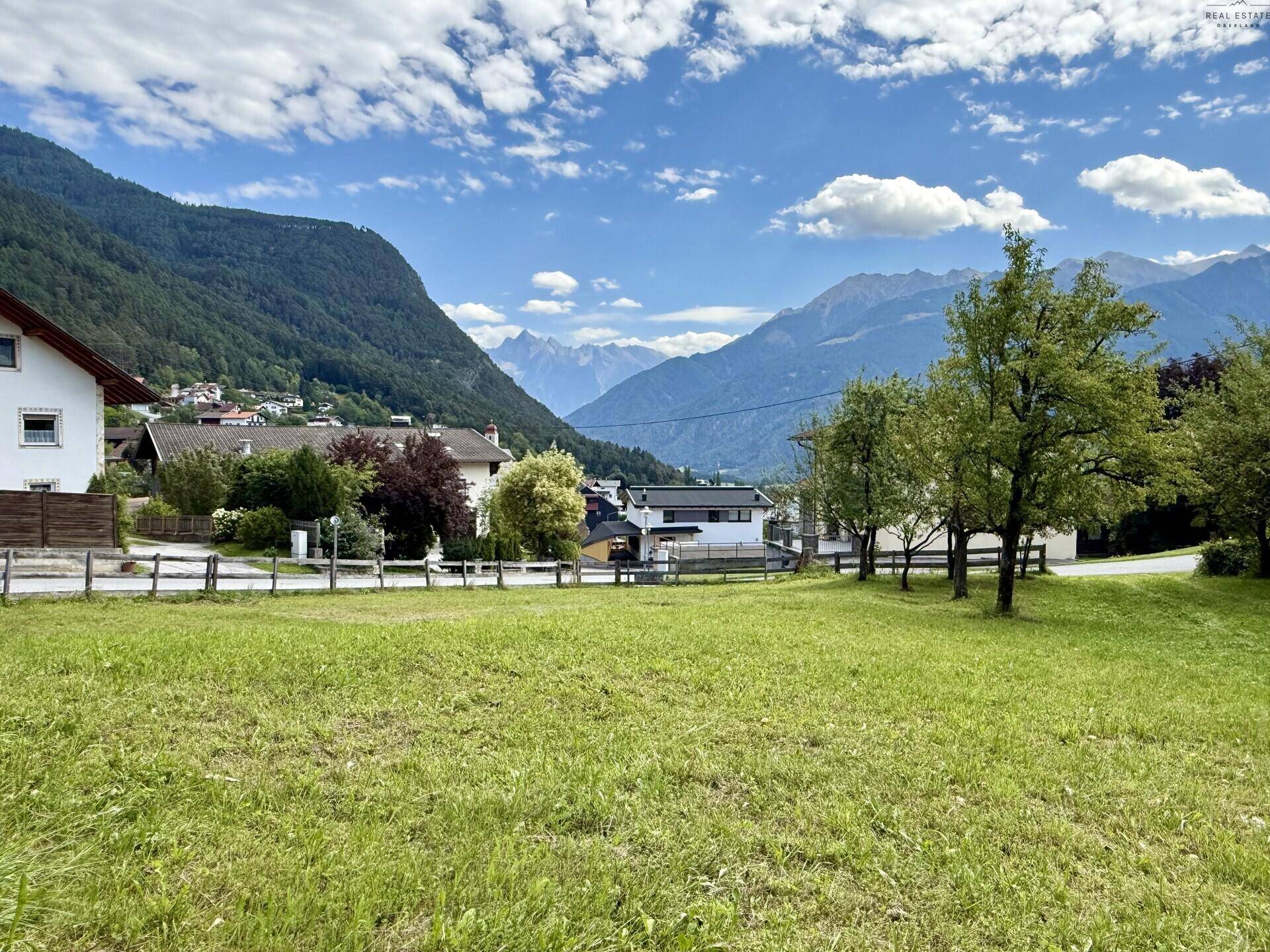 Panoramablick auf eine ländliche Siedlung mit grünen Hügeln und majestätischen Bergen im Hintergrund.