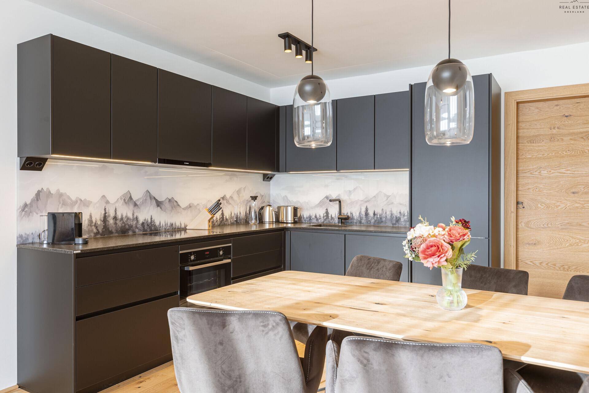 Moderne schwarze Küche mit integrierten Geräten, Holztisch und Bergpanorama-Rückwand.