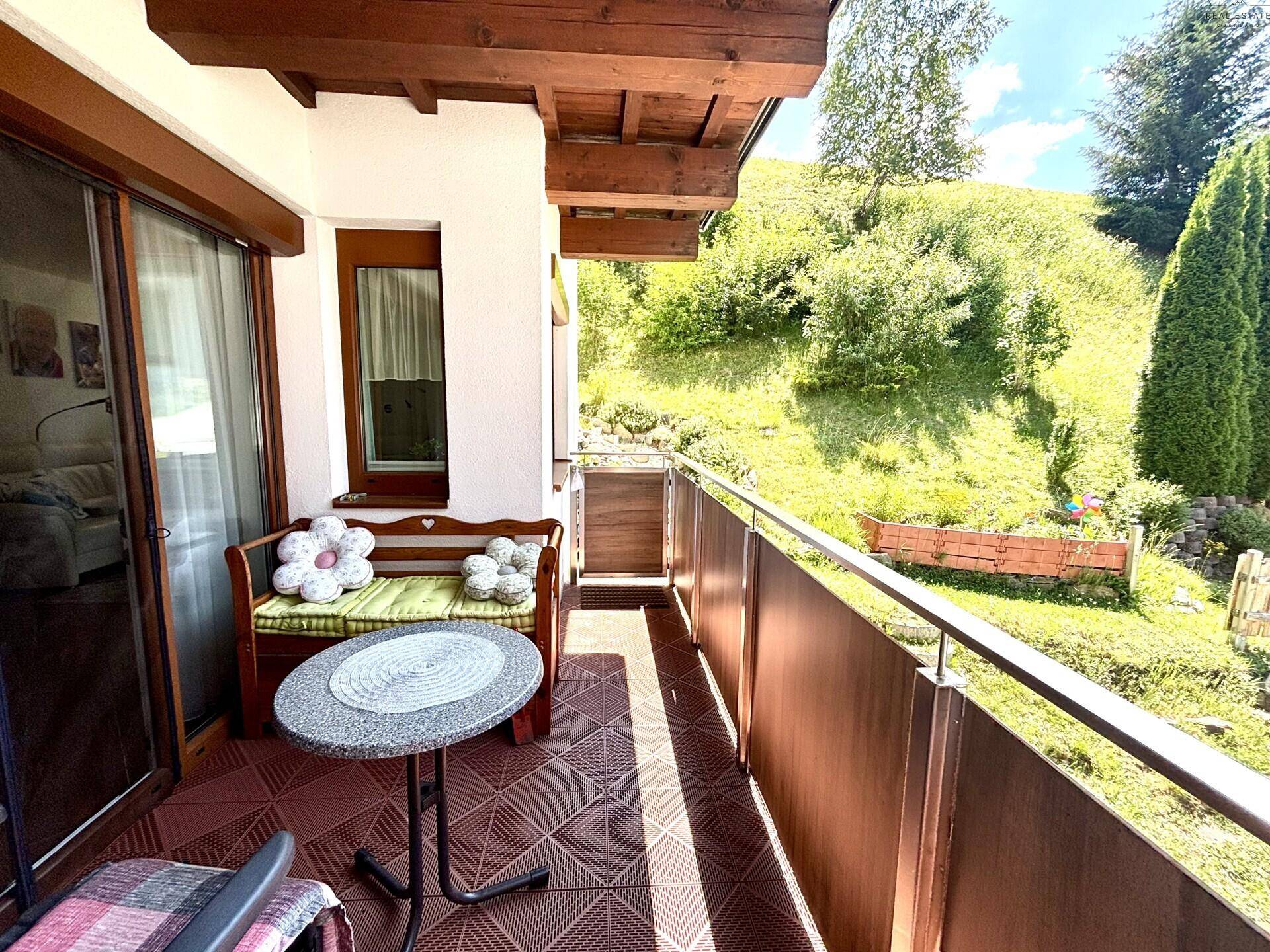 Sonniger Balkon mit Holzbalken, Sitzgelegenheiten und Blick auf die grüne Hanglandschaft.