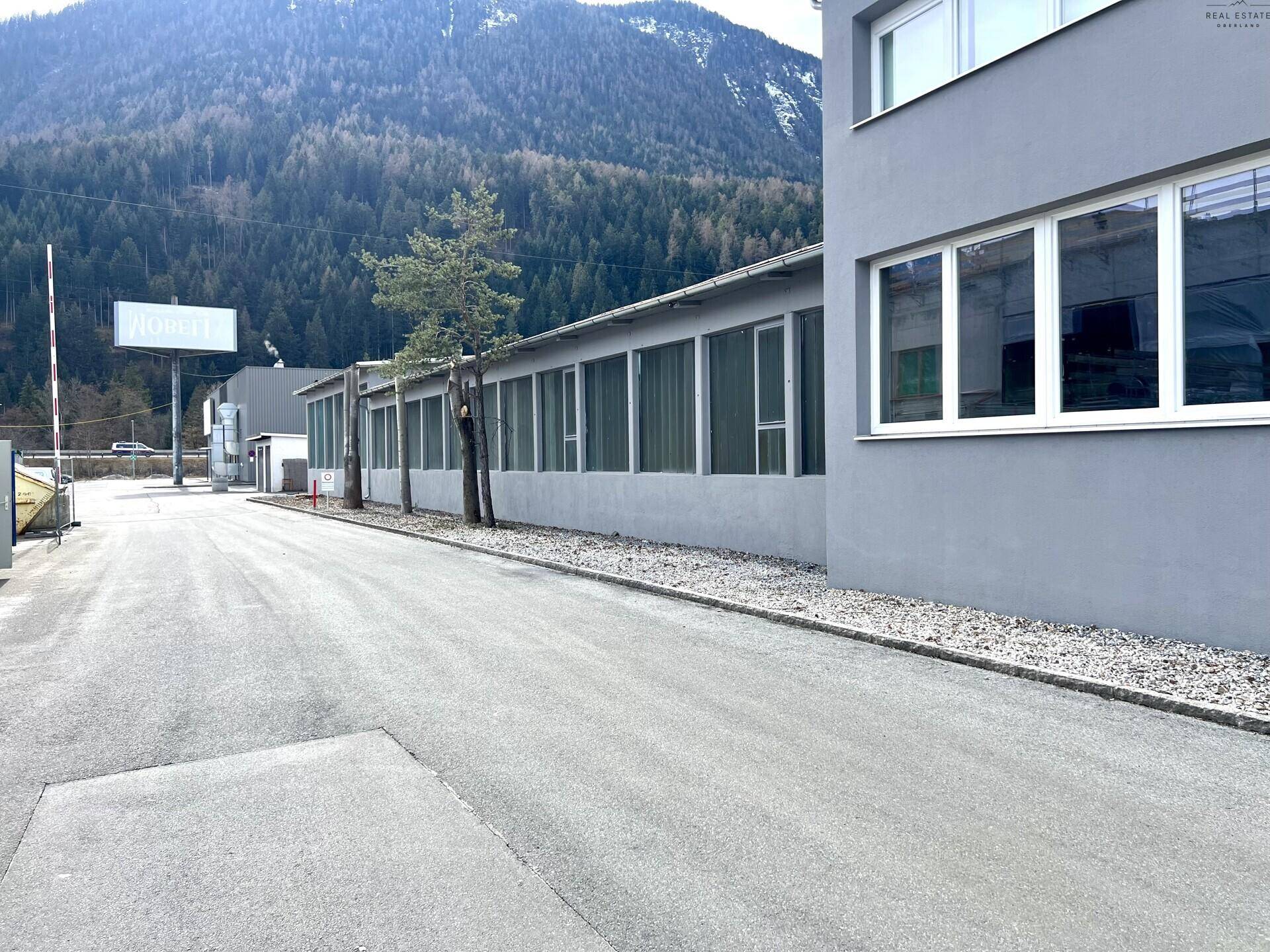 Seitenansicht des Industriegebäudes mit langen Fensterreihen, umgeben von Bäumen und Bergen.