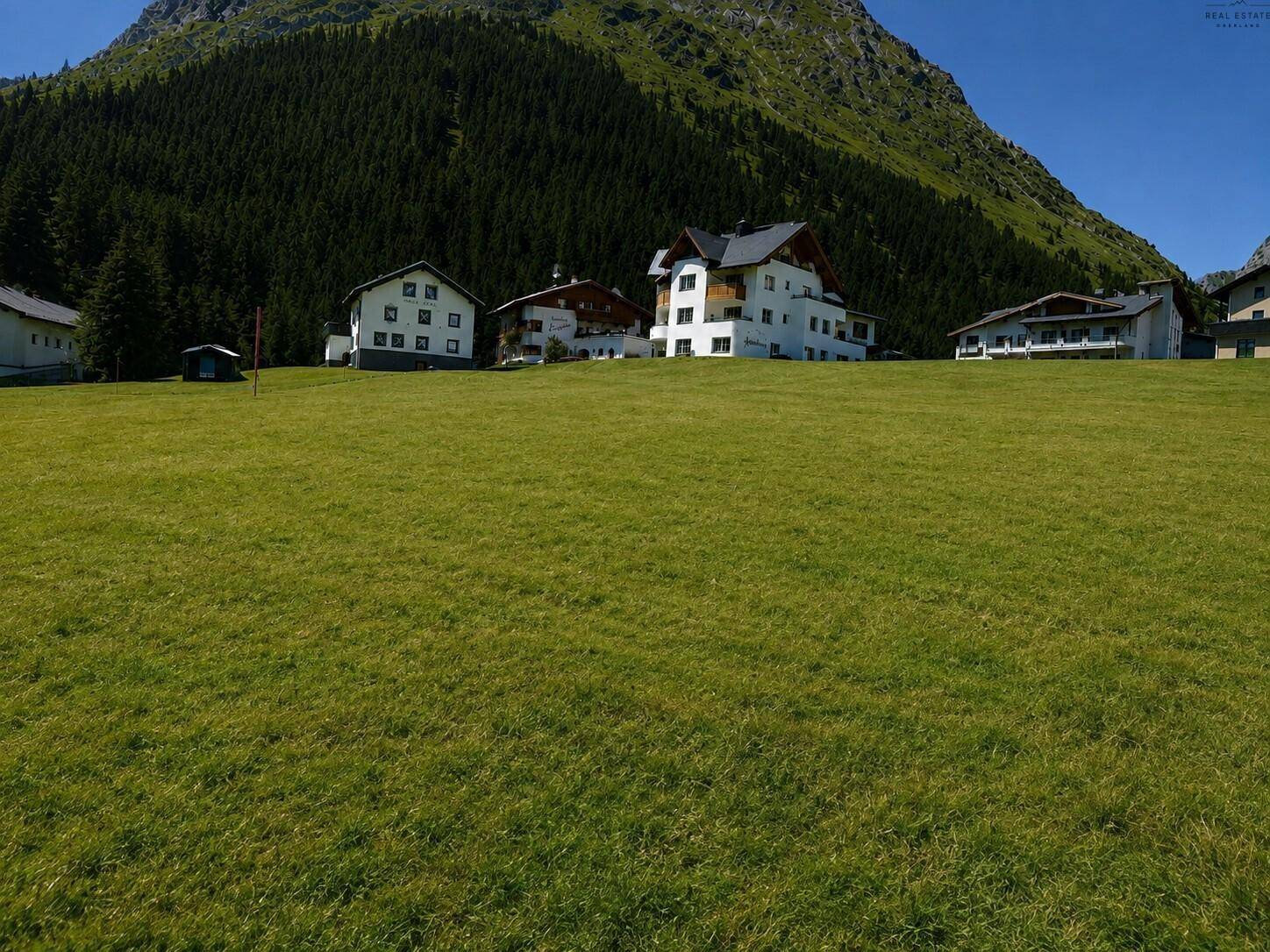 Weitläufige grüne Wiese mit traditionellen Gebäuden und majestätischem Bergpanorama im Hintergrund unter blauem Himmel.