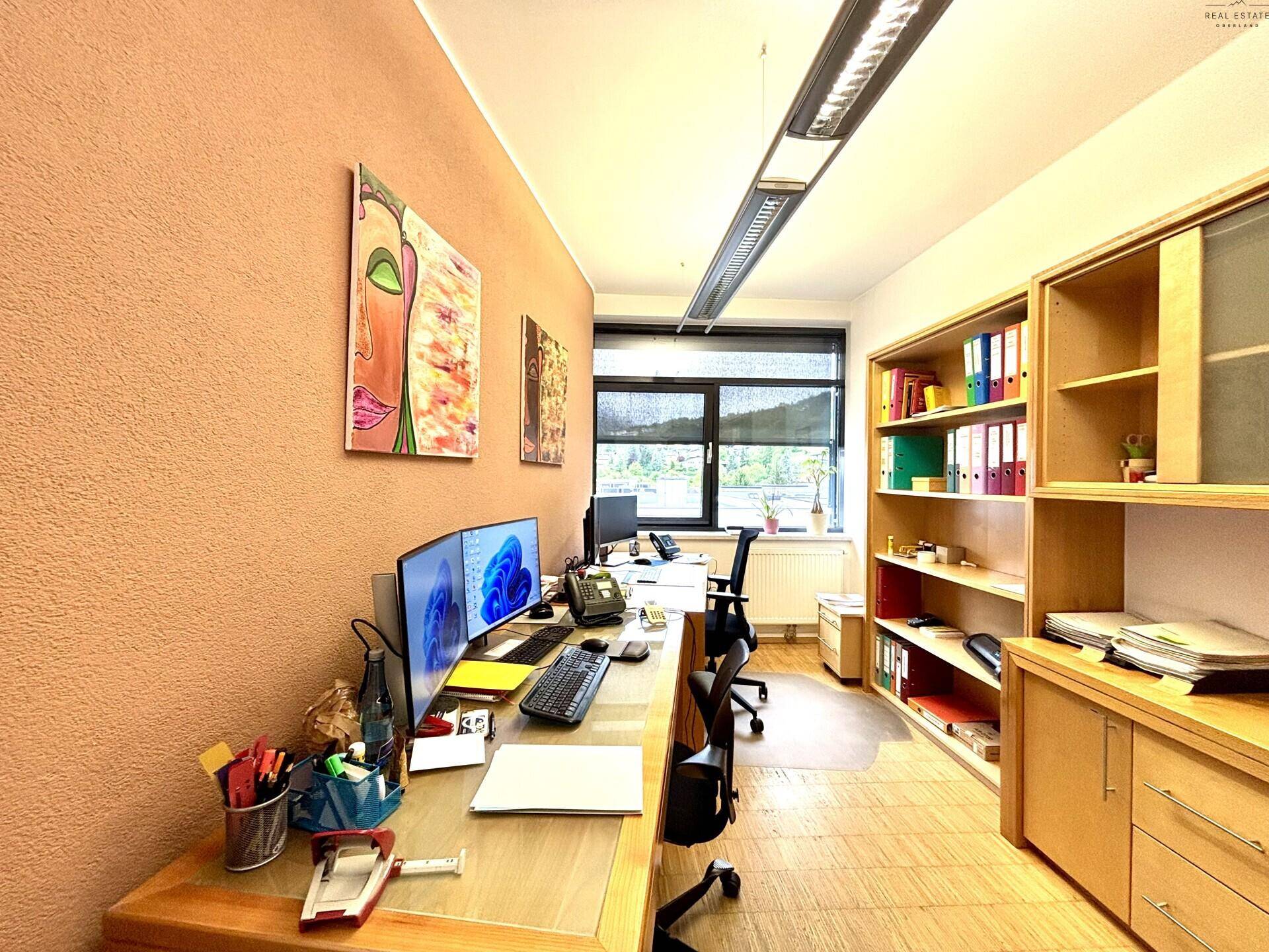 Bürozimmer mit zwei Arbeitsplätzen, Computern, Holzregalen und Fenstern mit Ausblick.