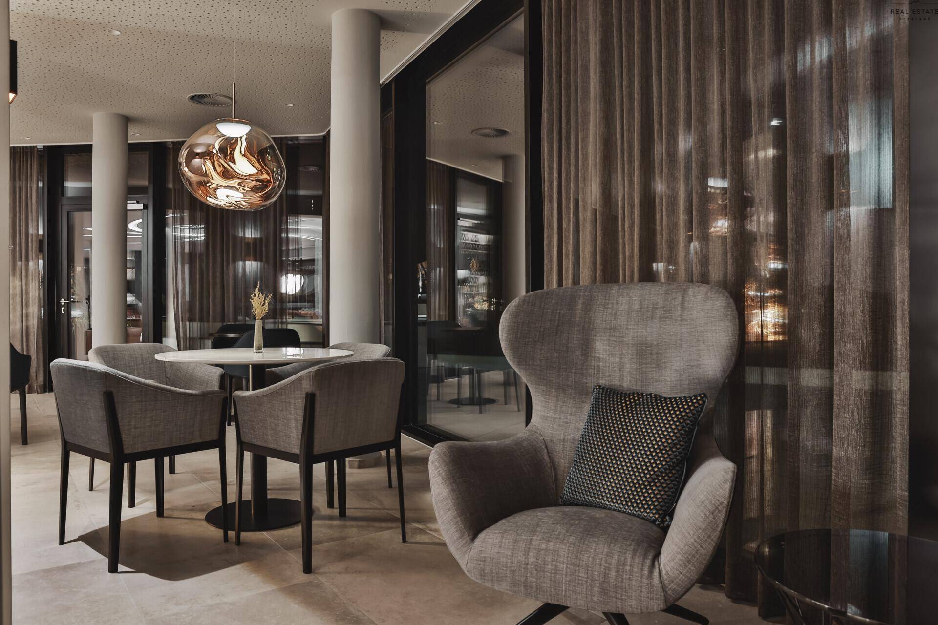 Elegante Lounge mit bequemen Sesseln, modernen Leuchten und großen Fenstern mit Vorhängen.