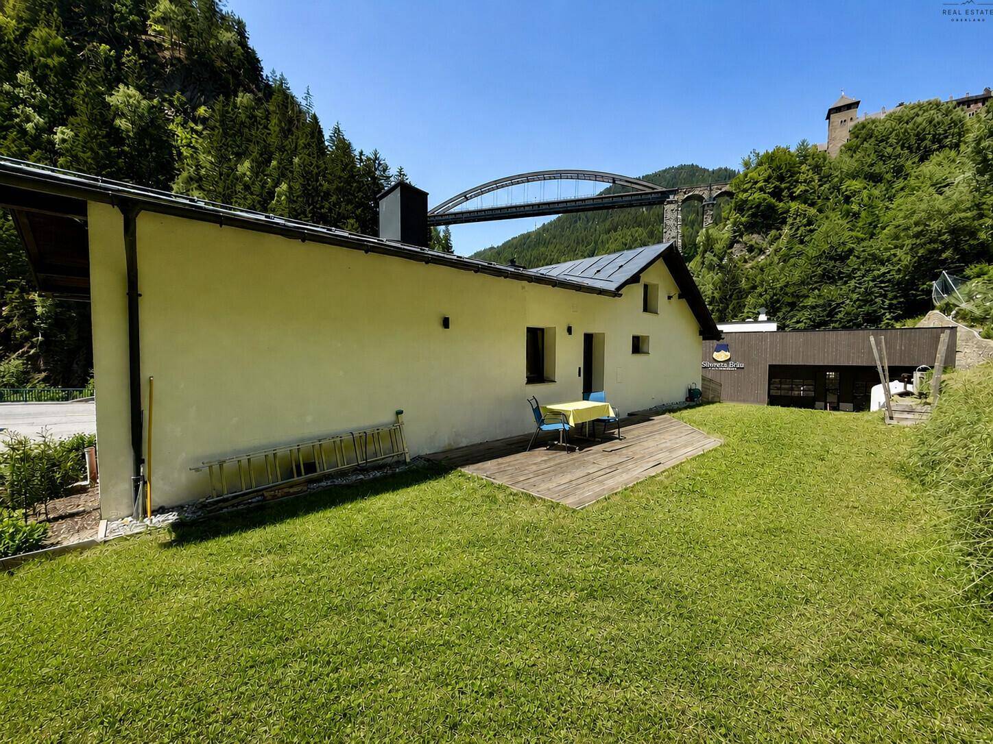 Gepflegter Rasenbereich mit kleiner Holzterrasse und Fernblick auf eine Brücke.