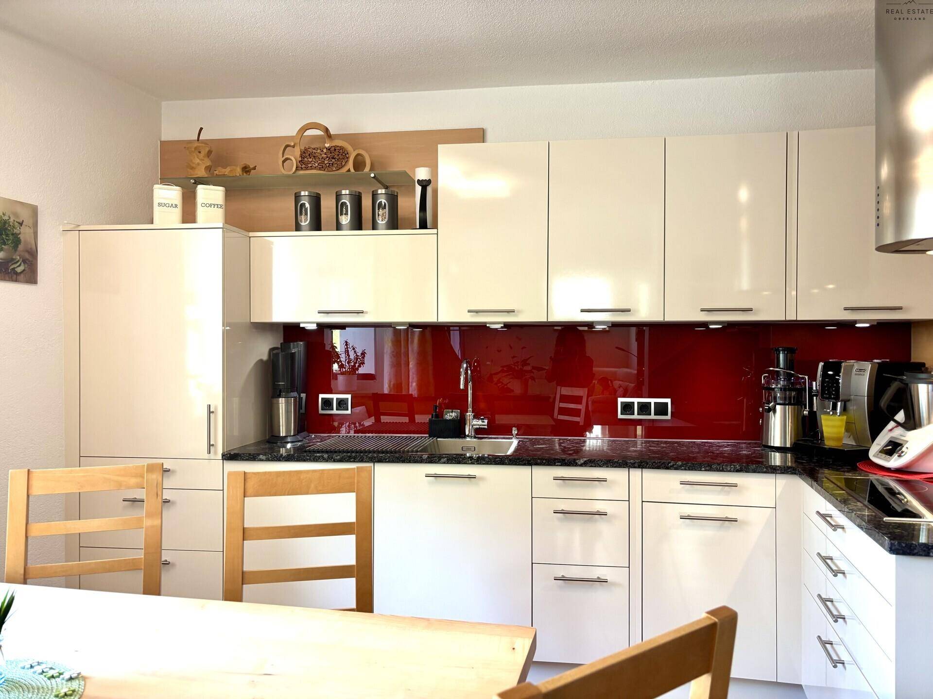 Moderne weiße Einbauküche mit glänzenden Fronten, dunkler Arbeitsplatte und roter Glasrückwand.