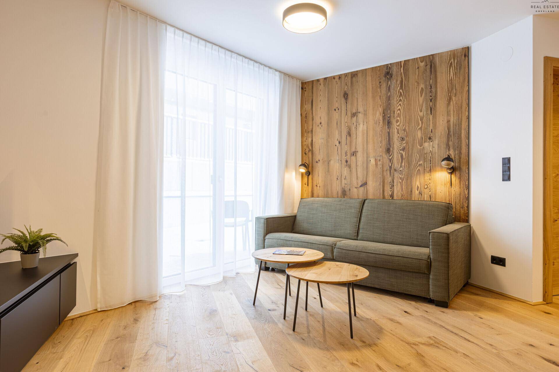 Helles Wohnzimmer mit Holzwand, beigem Sofa und Zugang zu einem Balkon oder einer Terrasse.