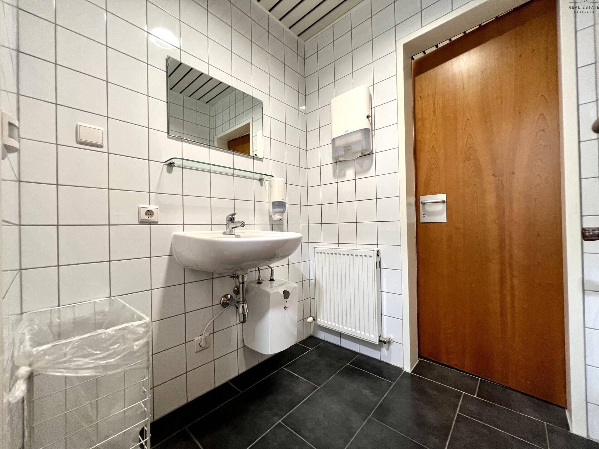 Helles Badezimmer mit weißen Fliesen, dunklem Boden und einem Waschbecken mit Spiegel.
