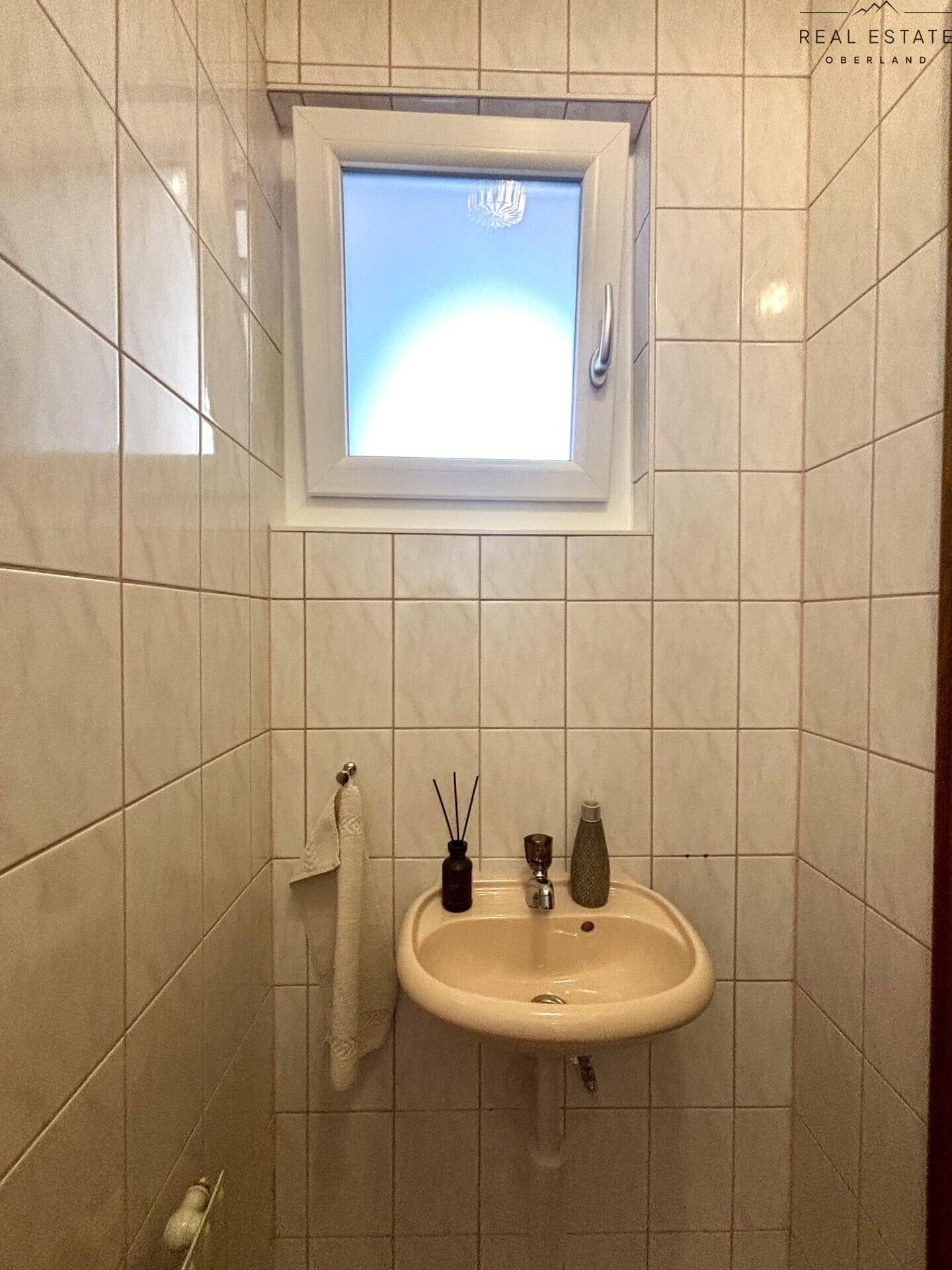 Kleines WC mit Fenster, beigen Fliesen und einem einzelnen Waschbecken.