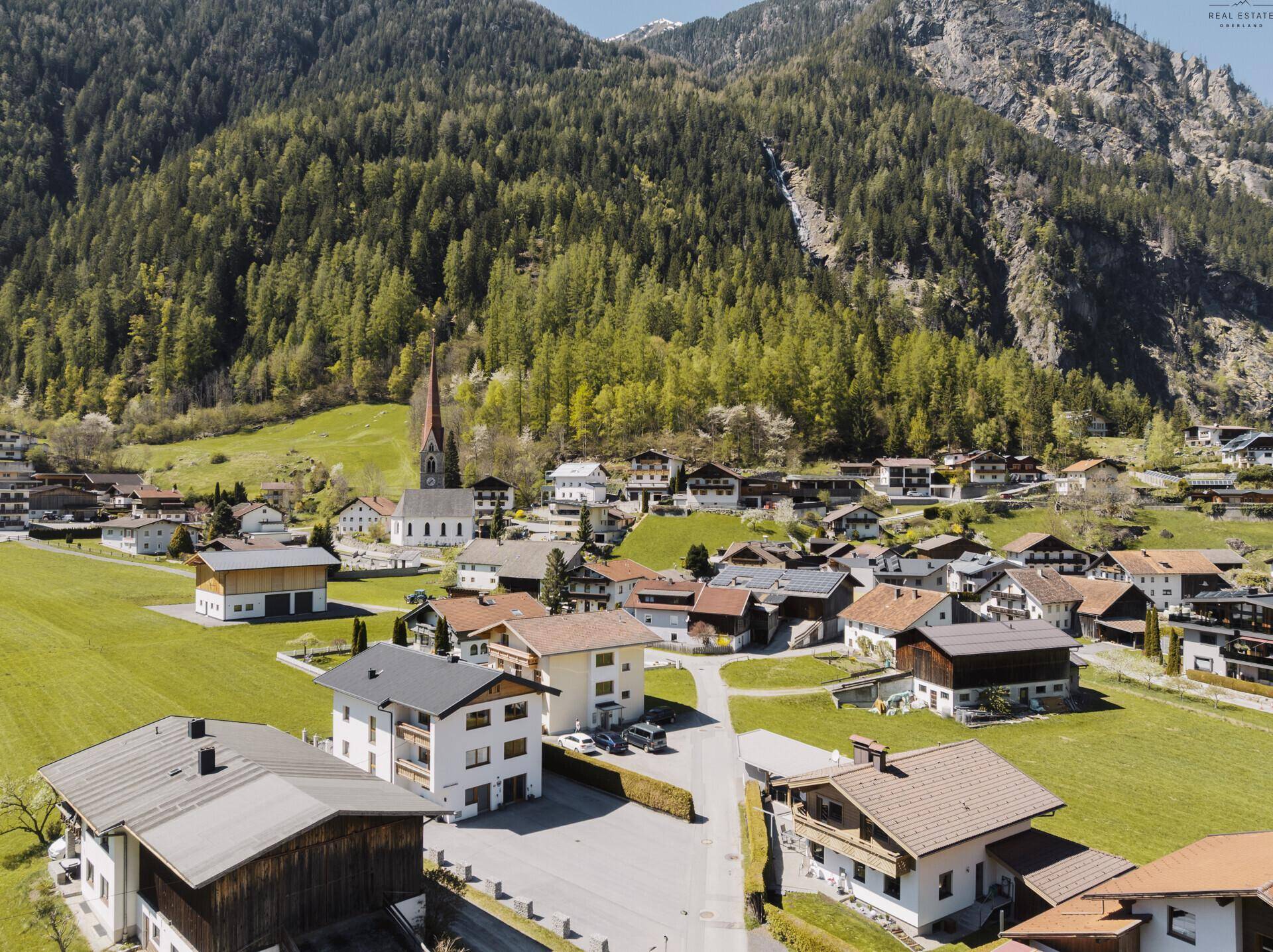 Luftaufnahme eines malerischen Dorfes in einem Tal, umgeben von grünen Bergen und Wäldern.