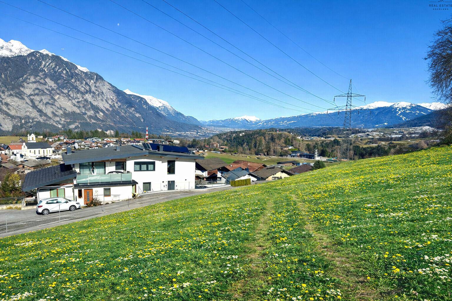 Weitblick über eine grüne Wiese mit Häusern im Tal und schneebedeckten Gipfeln am Horizont.