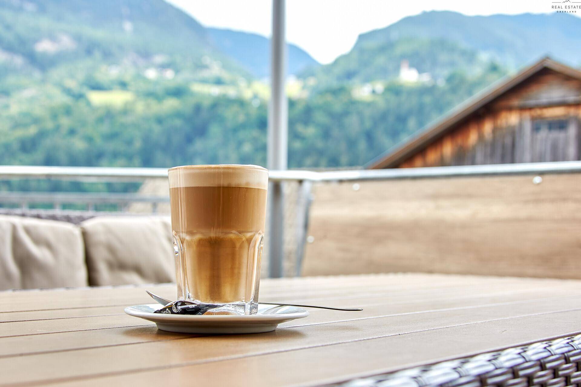 Nahaufnahme eines Kaffees auf einem Holztisch mit Rattanmöbeln und Bergblick im Hintergrund.