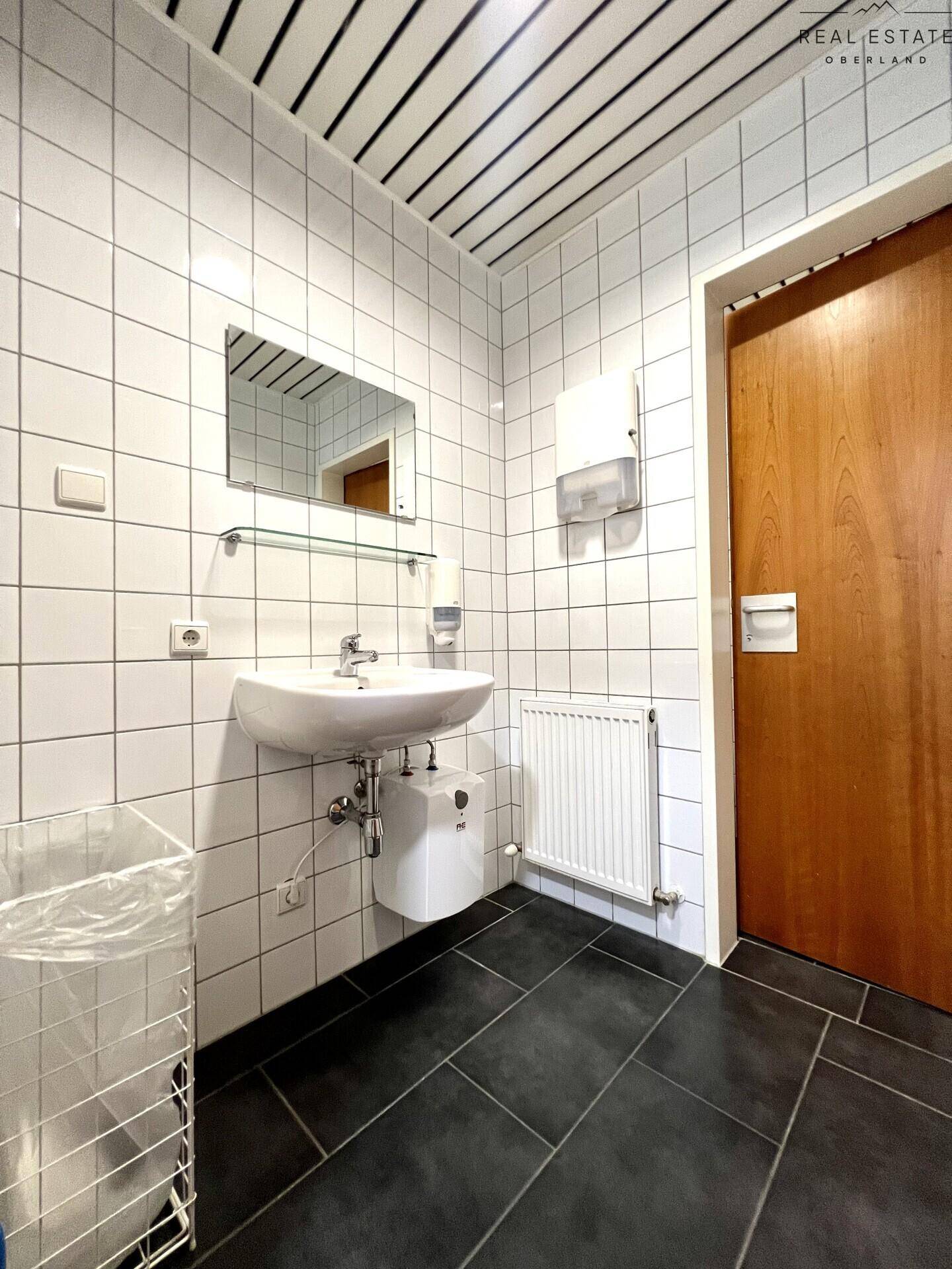 Sauberes Badezimmer mit weißen Wandfliesen, dunklem Boden und einem Waschbecken mit Spiegel.
