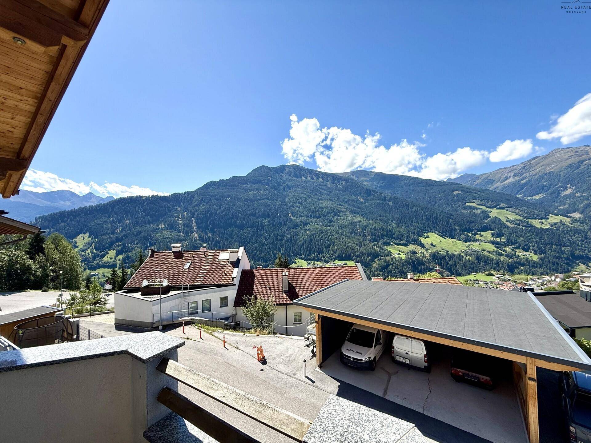 Balkon mit weitem Blick auf die umliegende Berglandschaft und ein Carport im Vordergrund.