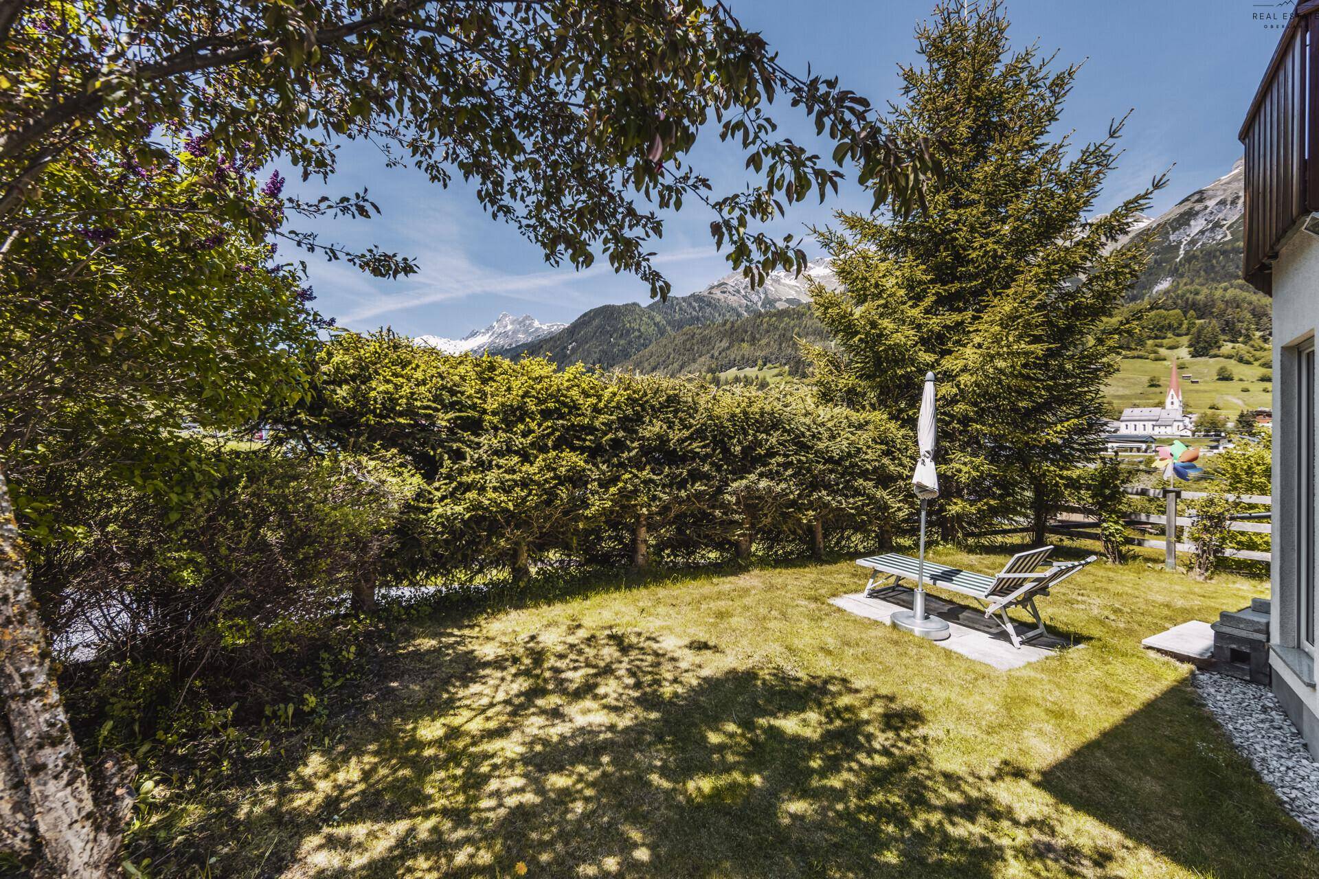 Gepflegter Gartenbereich mit Liegestuhl, Hecke und Bergblick unter blauem Himmel.