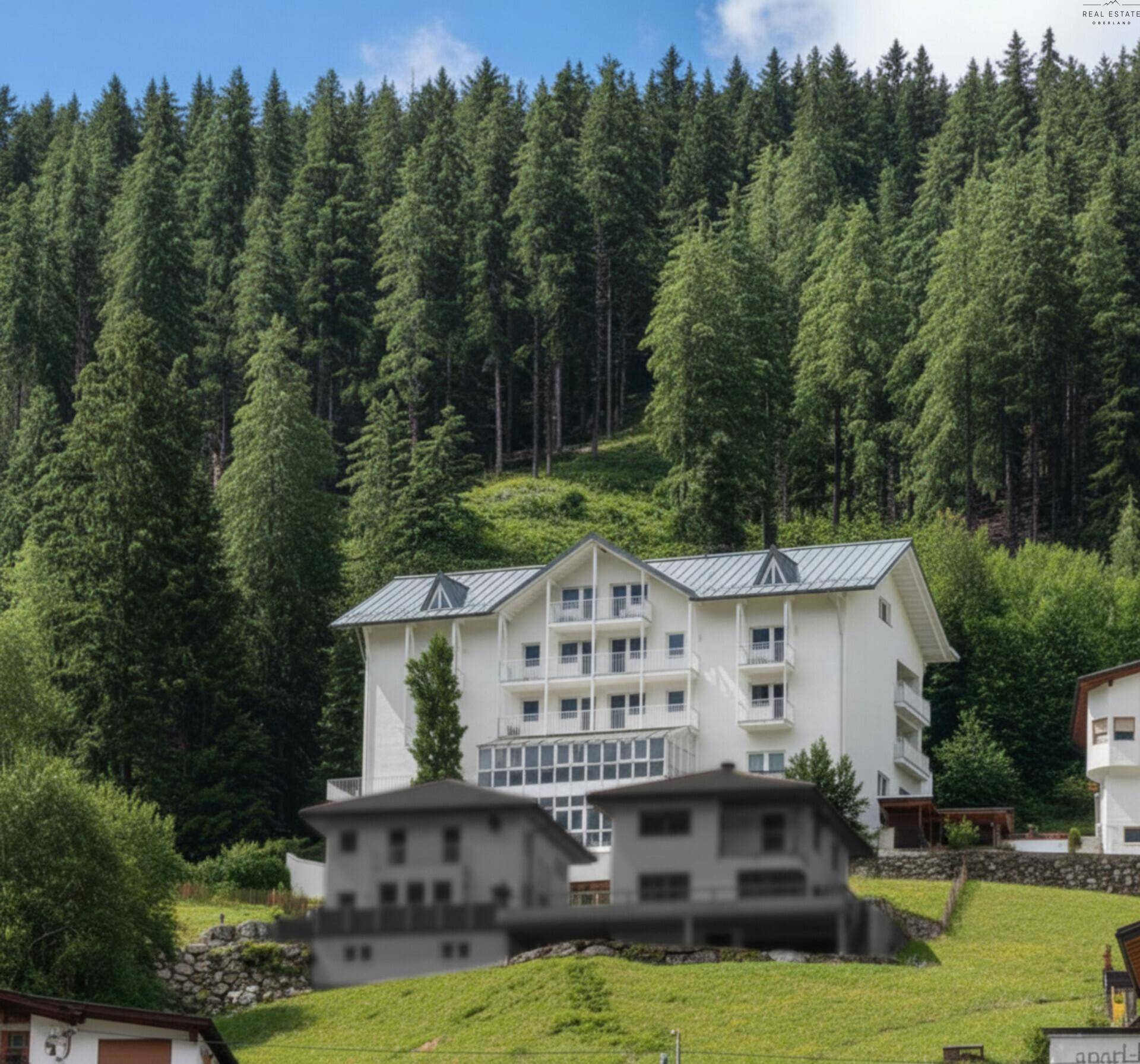 Weißes Gebäude in alpiner Landschaft, umgeben von hohen Nadelbäumen unter blauem Himmel.