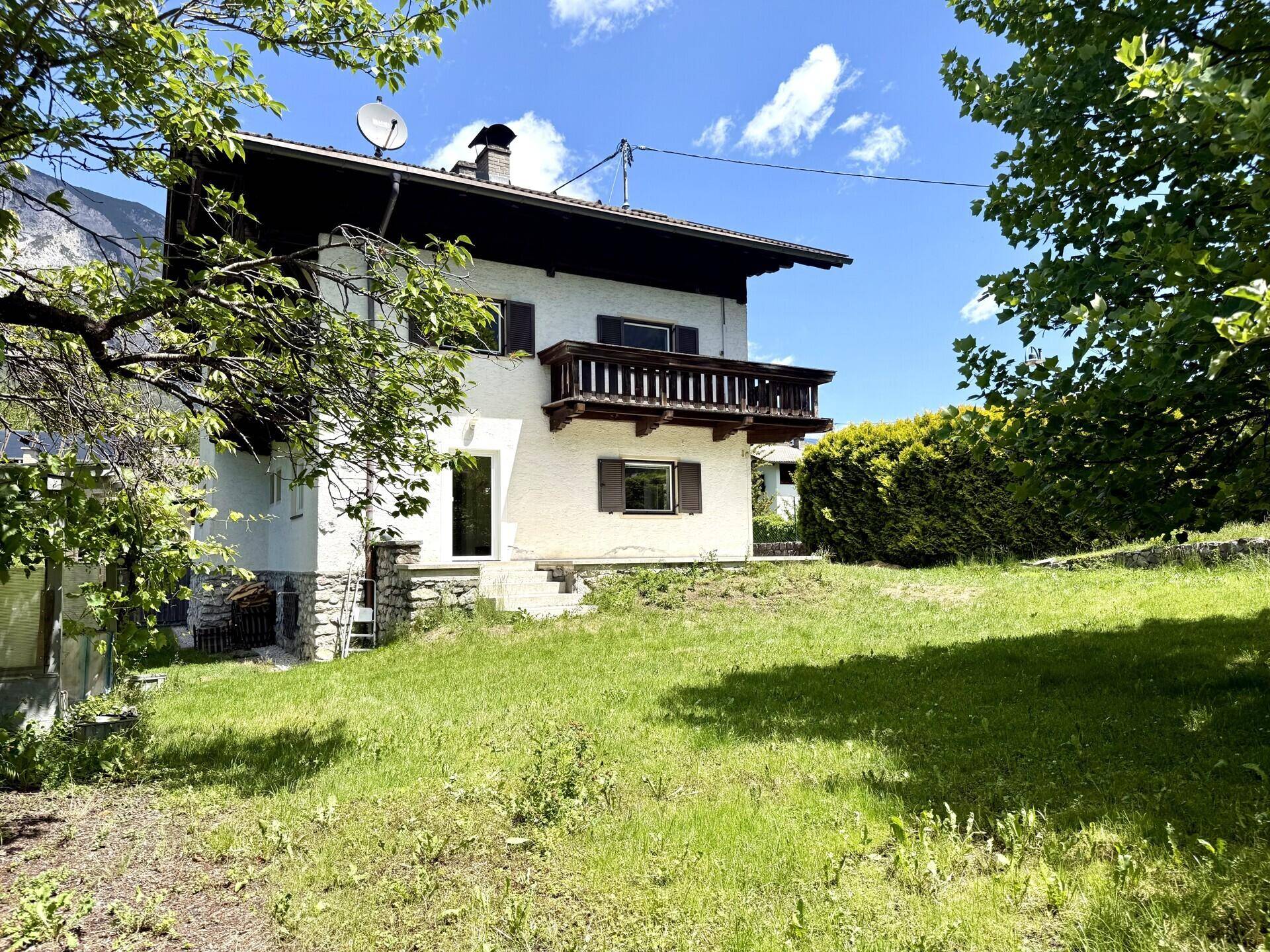 Haus mit Holzbalkon und großem Garten, umgeben von Bäumen und Bergen.