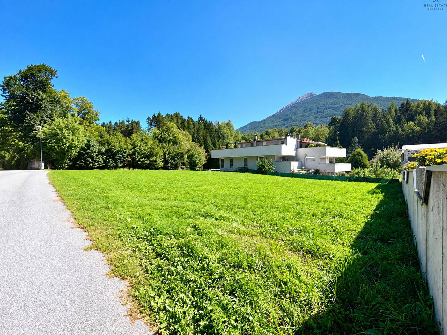 Weitläufige Grünfläche mit angrenzendem Wald und einem Haus im Hintergrund.