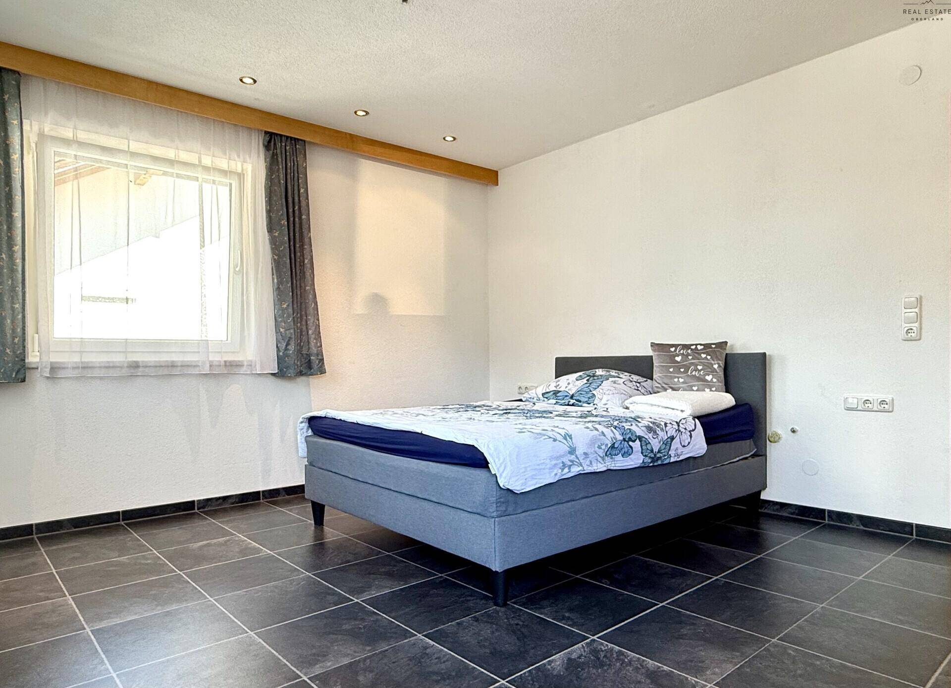 Helles Schlafzimmer mit dunklem Fliesenboden, Doppelbett und einem Fenster mit Tageslicht.