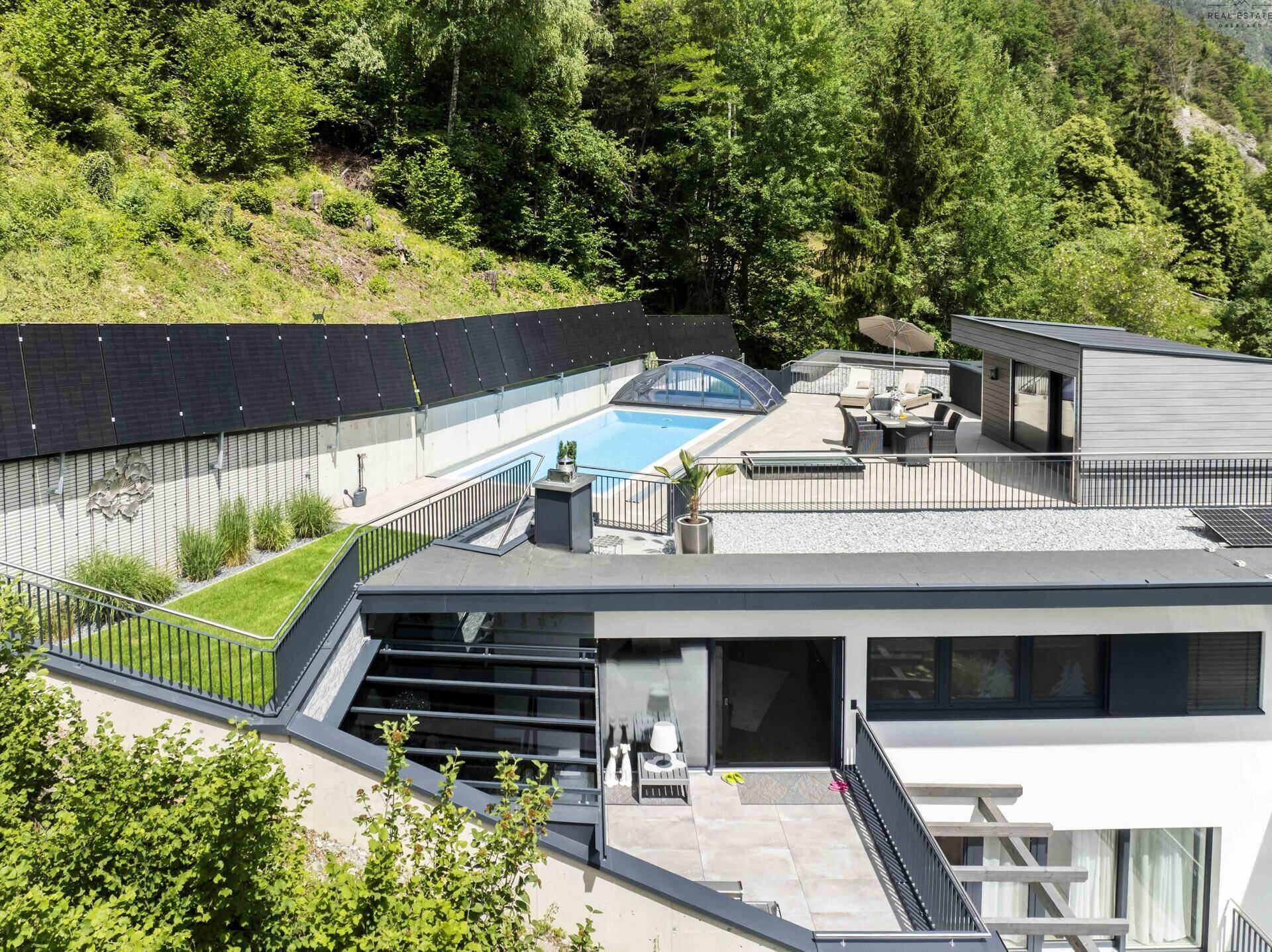 Luftaufnahme des modernen Anwesens mit Pool, großer Terrasse und integrierten Solarpaneelen in grüner Umgebung.