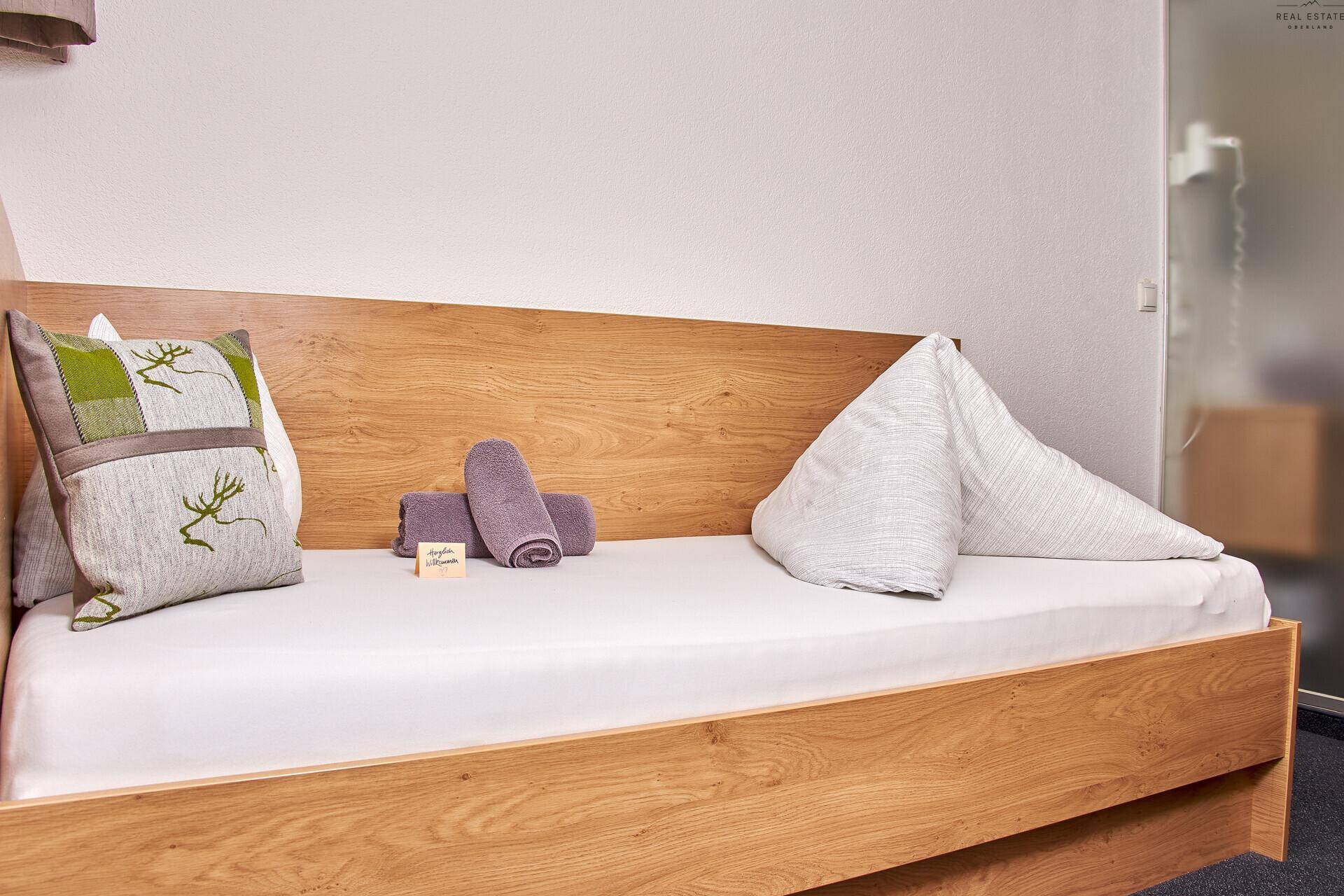 Einzelbett mit Holzbettrahmen und grünen Kissen in einem hellen, einfach eingerichteten Zimmer.