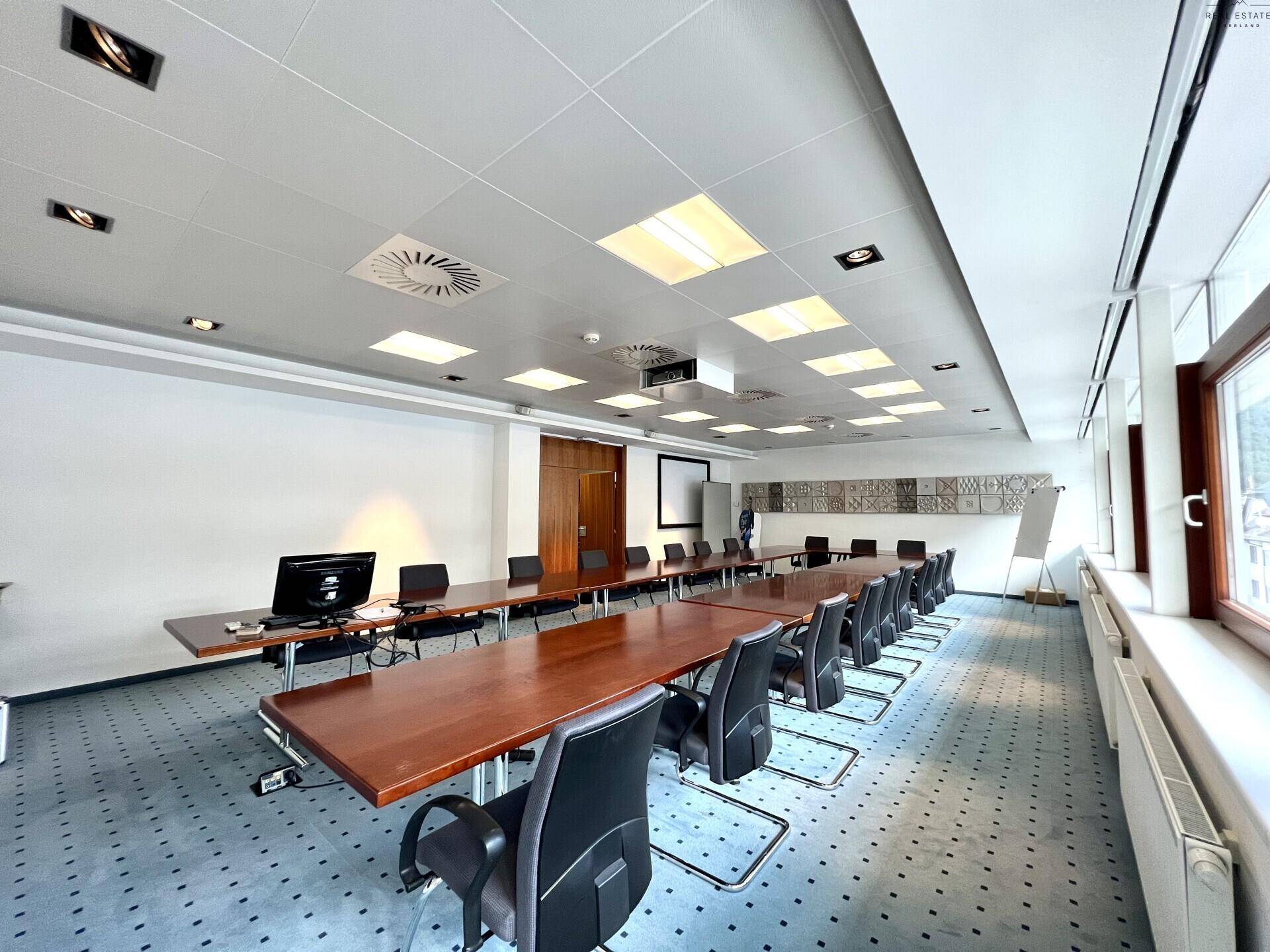 Heller Konferenzraum mit mehreren Tischen, Bürostühlen und einem Projektor für Präsentationen.