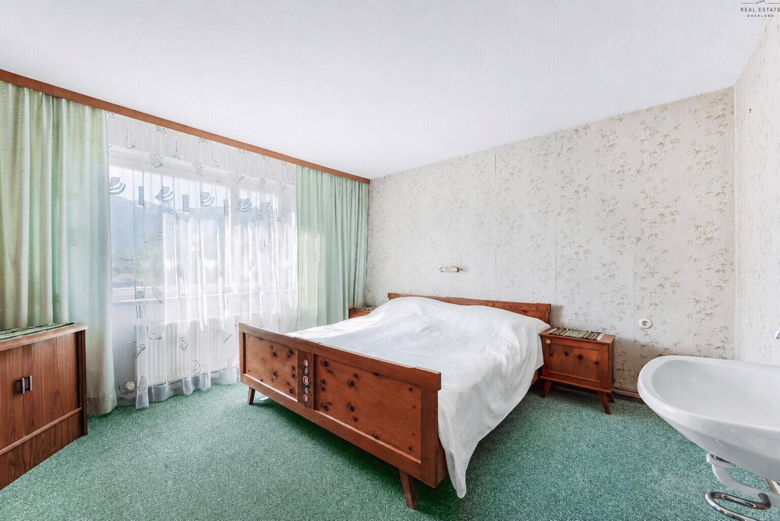 Schlafzimmer mit Doppelbett, Holzmöbeln und einem Waschbecken, hell durch große Fenster.