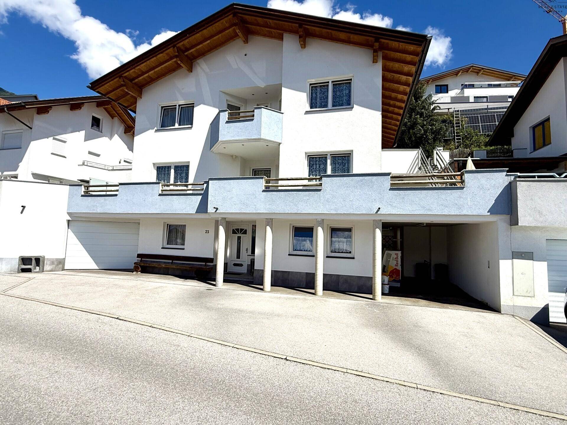Mehrstöckiges weißes Haus mit blauen Balkonen, großem Vorplatz und Carport in bergiger Umgebung.