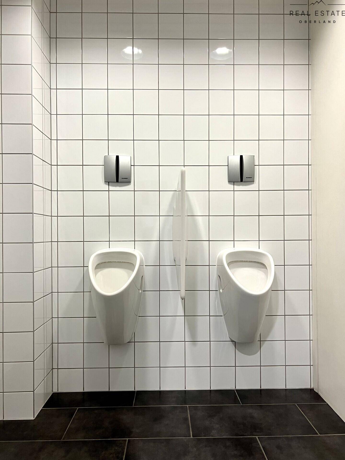Herren-WC mit zwei Urinalen, weißen Fliesen und dunklem Boden in einem gepflegten Zustand.