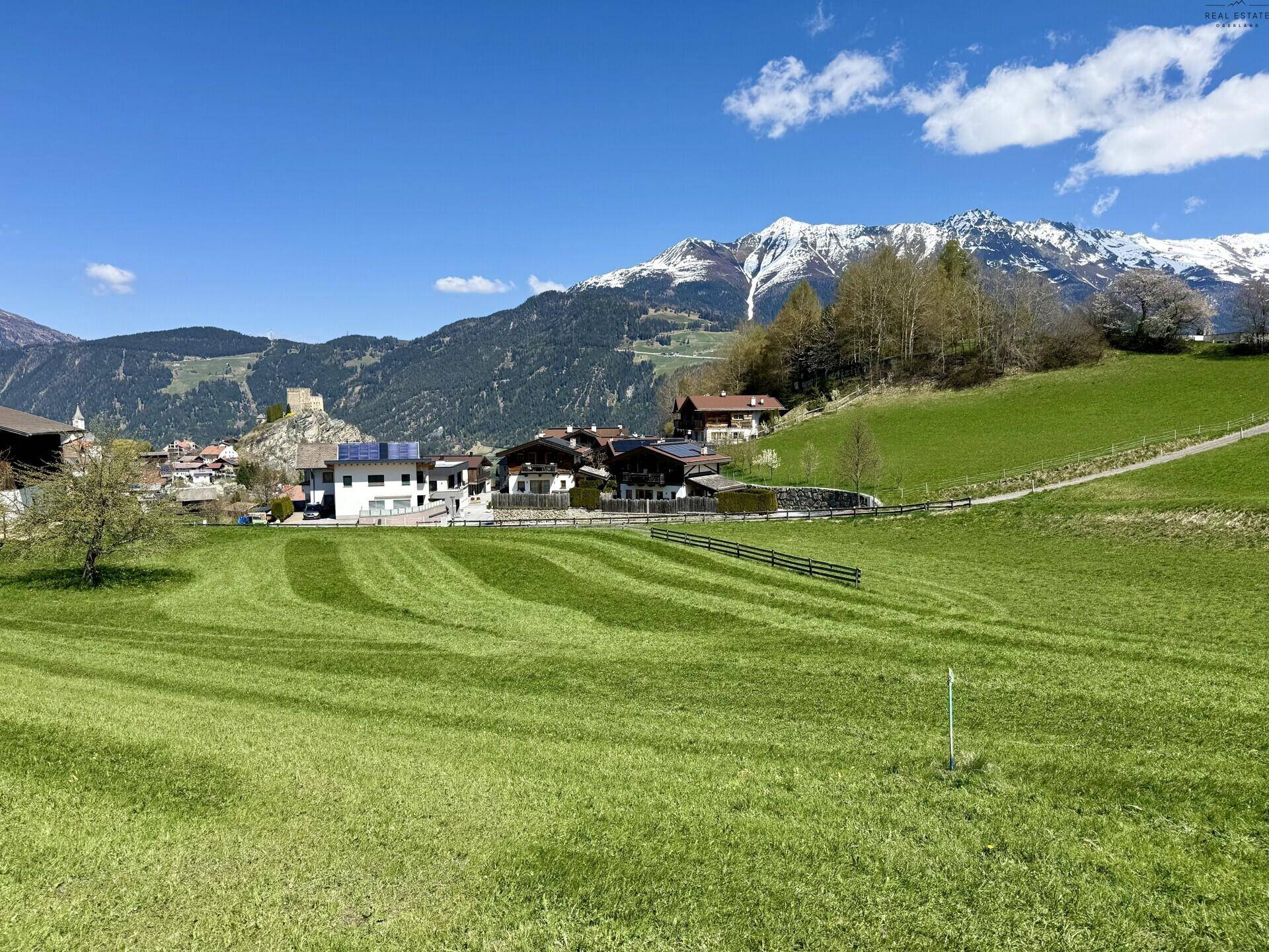 Gepflegte grüne Wiesen mit Blick auf ein historisches Dorf mit Burgruine und die majestätischen Alpen im Hintergrund.