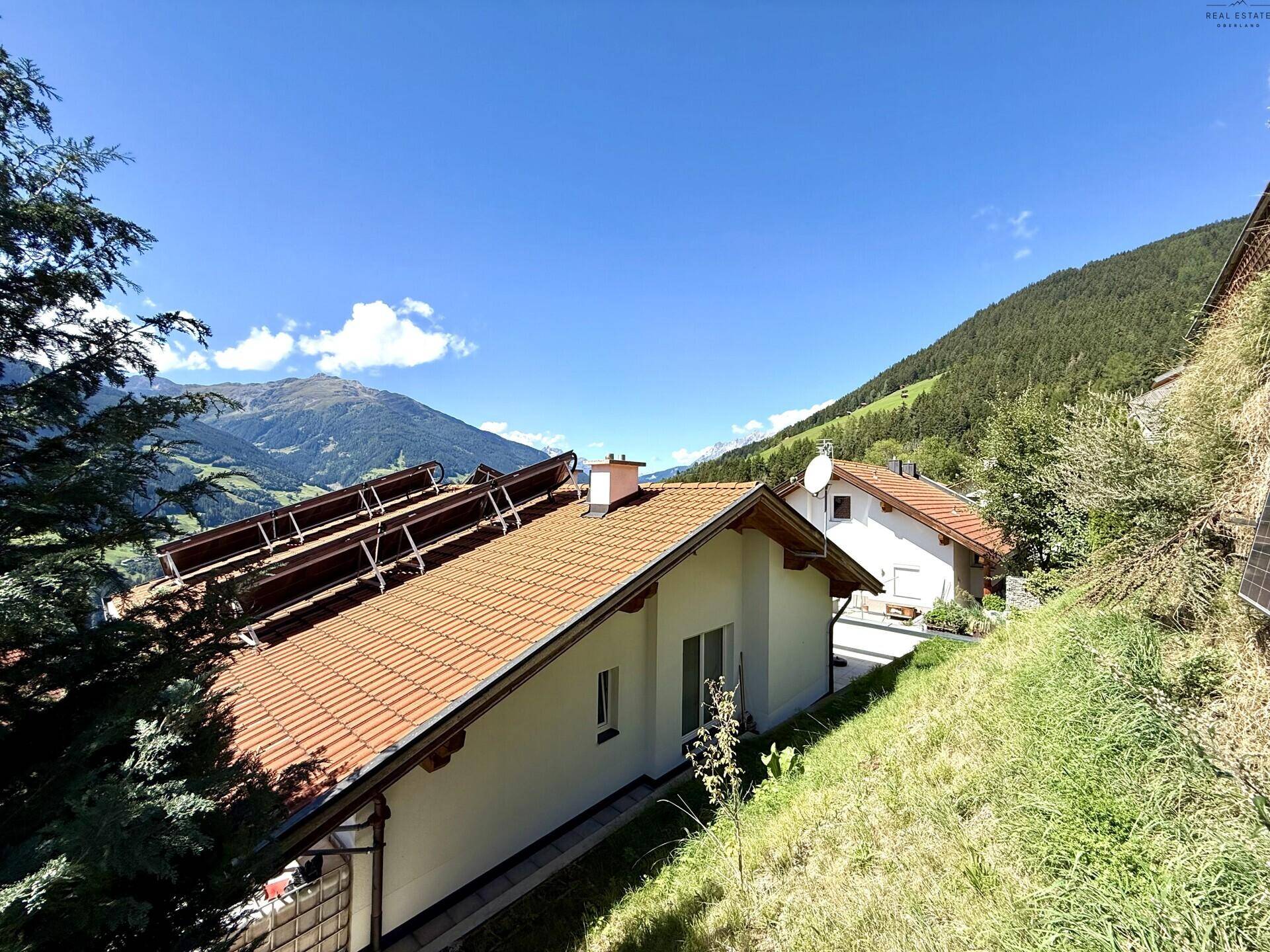 Haus auf einem Hügel mit Ziegeldach und Solaranlage, umgeben von Bergen und grüner Landschaft.