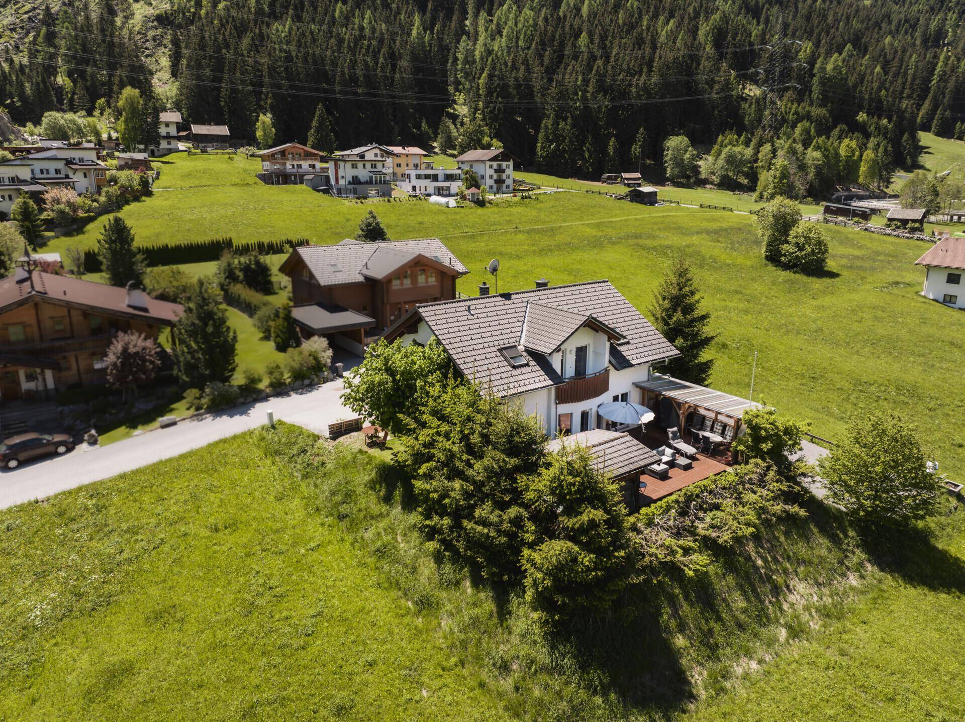 Luftaufnahme des Hauses mit gepflegtem Garten und einer großen Holzterrasse in alpiner Umgebung.