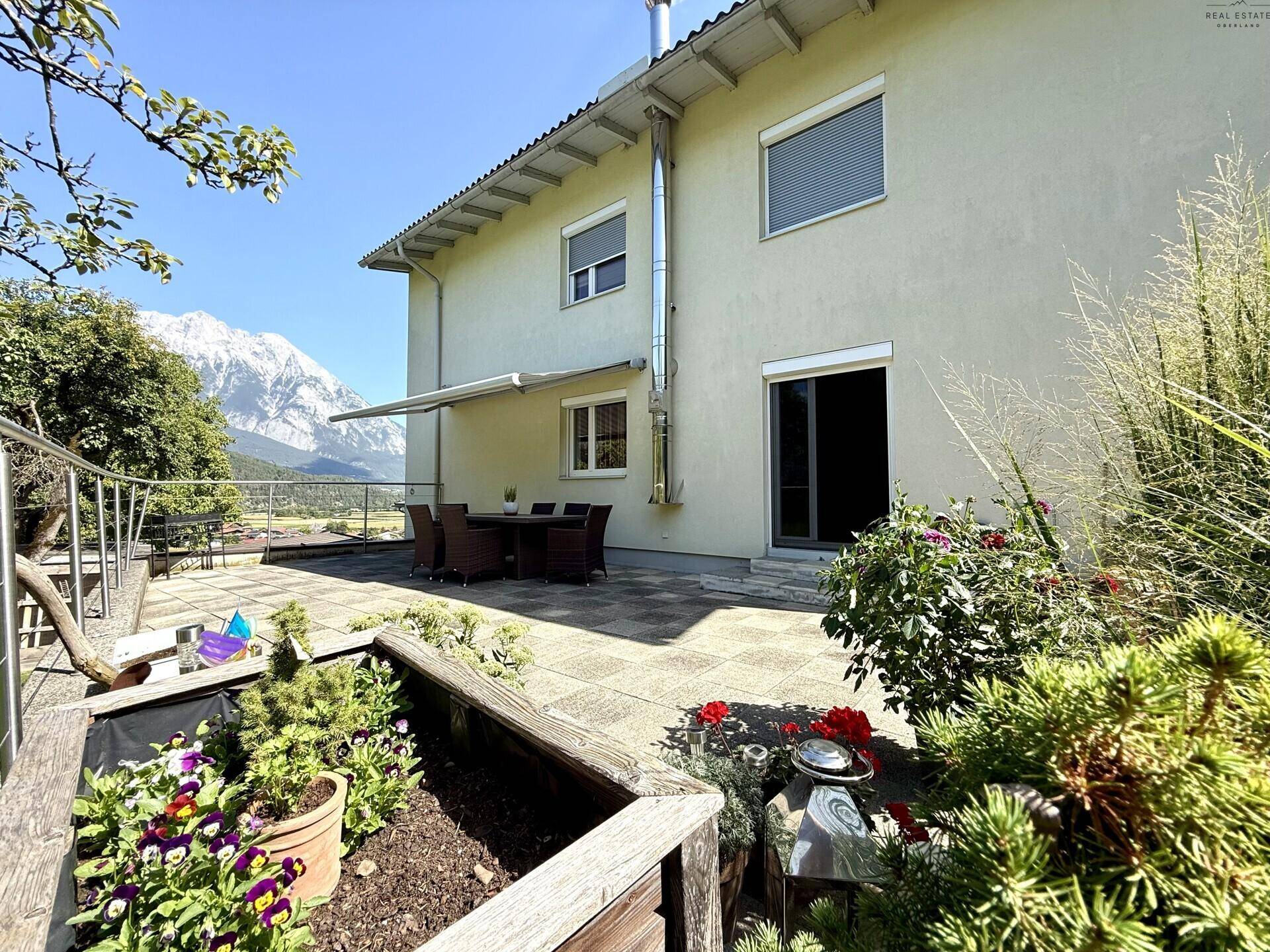 Hausfassade mit großer Terrasse, Gartenbeeten und beeindruckendem Bergblick in der Ferne.
