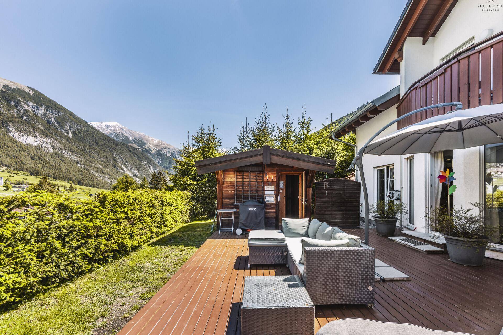 Sonnige Holzterrasse mit Lounge-Möbeln, umgeben von einer grünen Hecke und Bergblick.