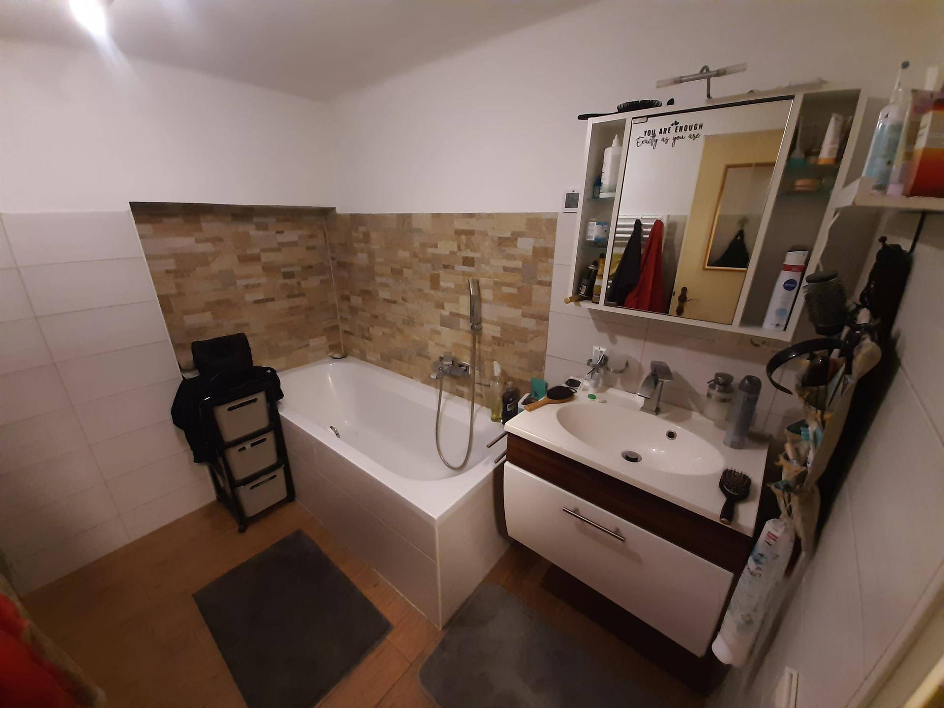 Helles Badezimmer mit Badewanne, Einzelwaschbecken und Spiegelschrank, akzentuiert durch eine Steinwand.