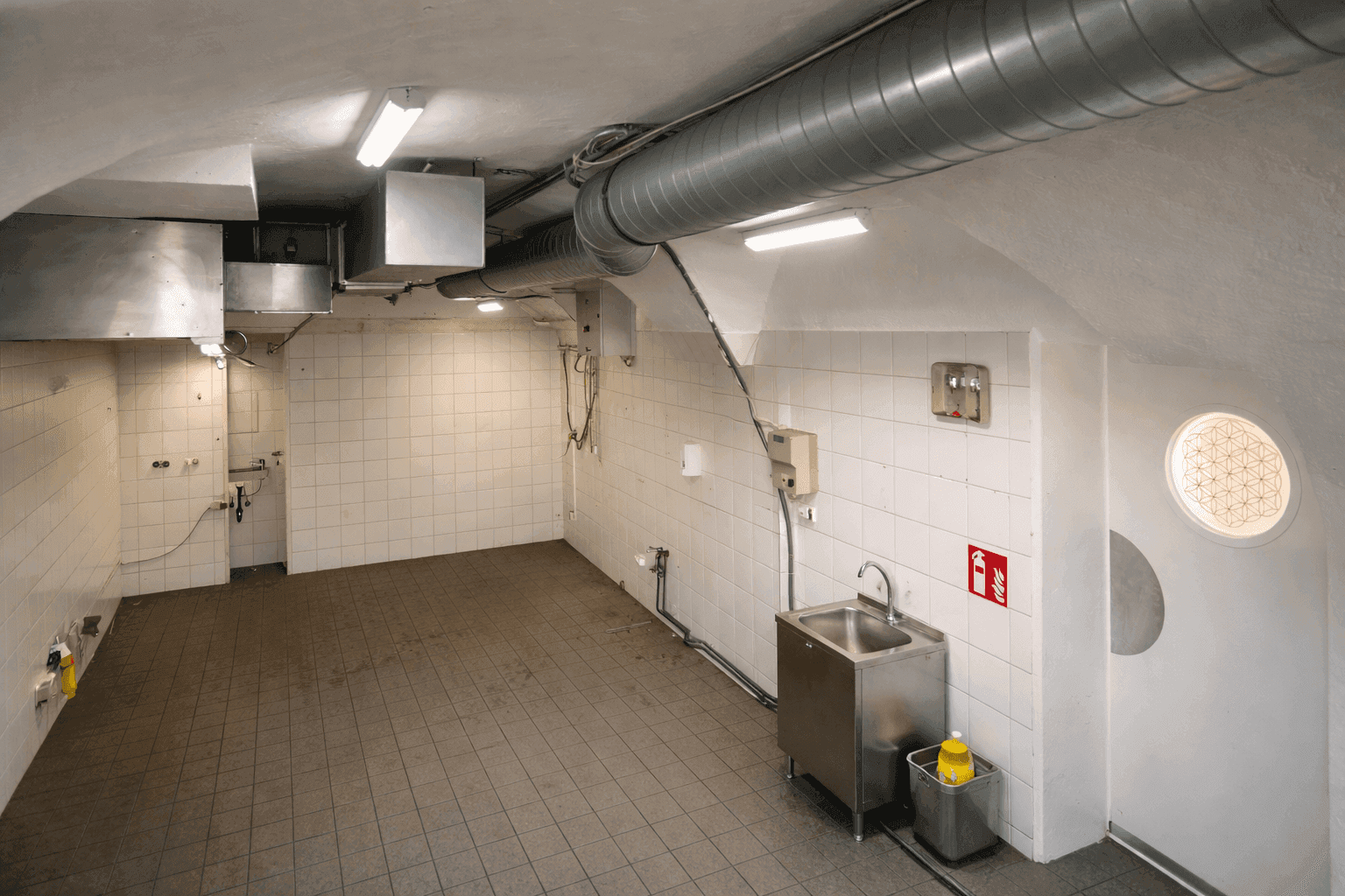 Geräumige Gewerbeküche mit gefliesten Wänden und Böden, Edelstahlspüle und sichtbaren Lüftungsrohren.
