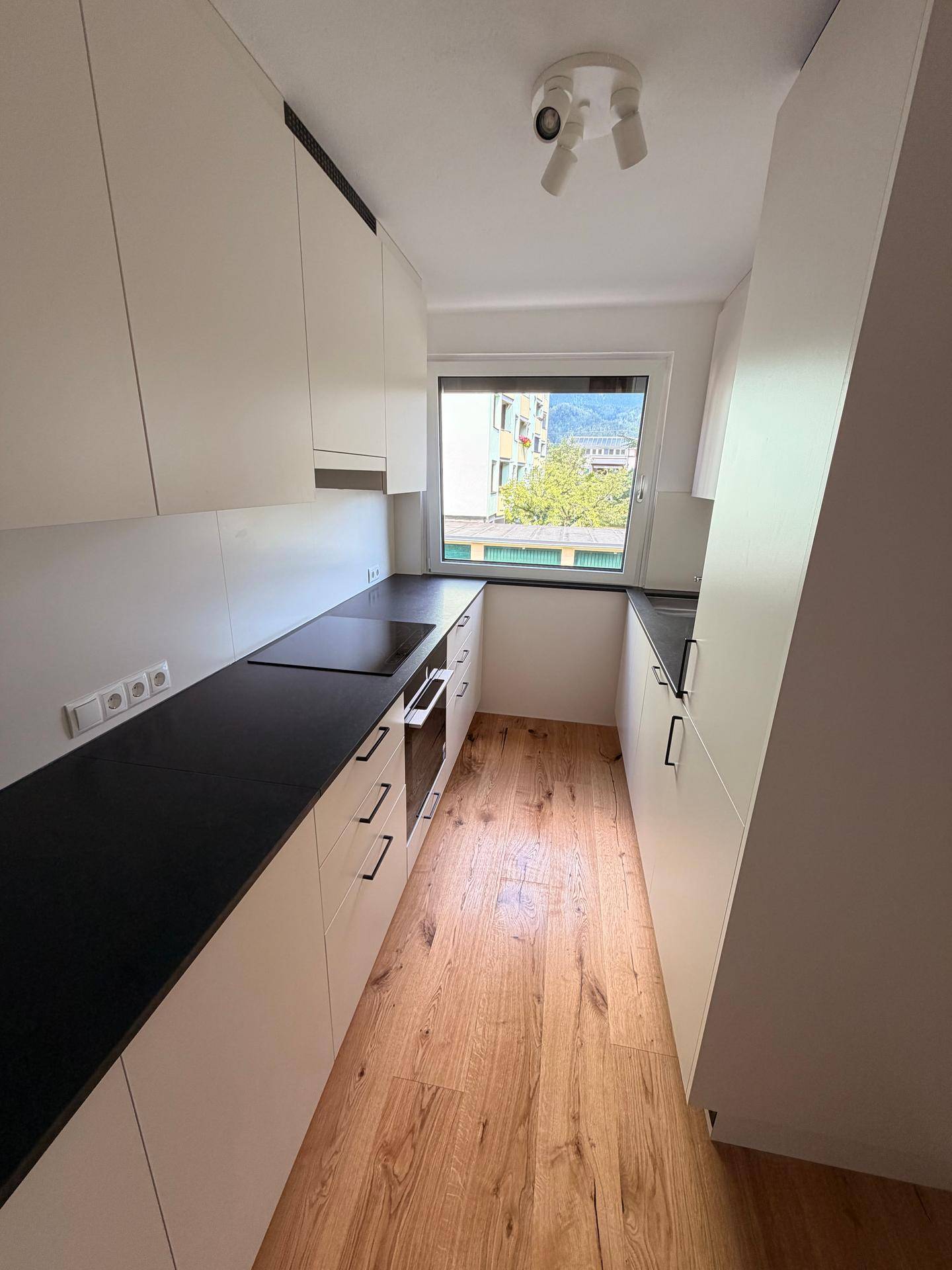 Moderne, schmale Küche mit weißen Schränken, dunkler Arbeitsplatte und hellem Holzfußboden.