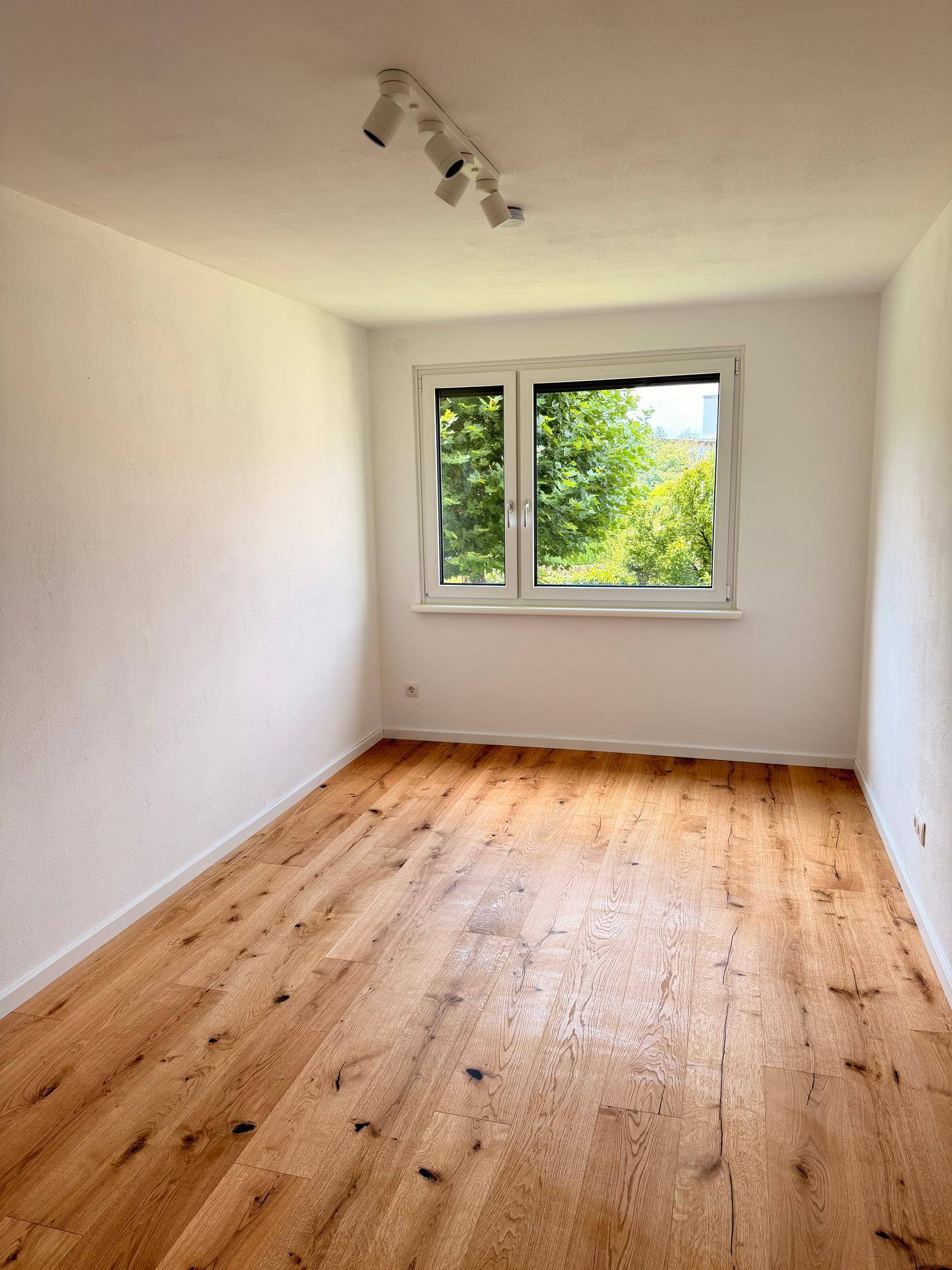 Helles Zimmer mit neuem Holzfußboden und Blick ins Grüne durch das Fenster.