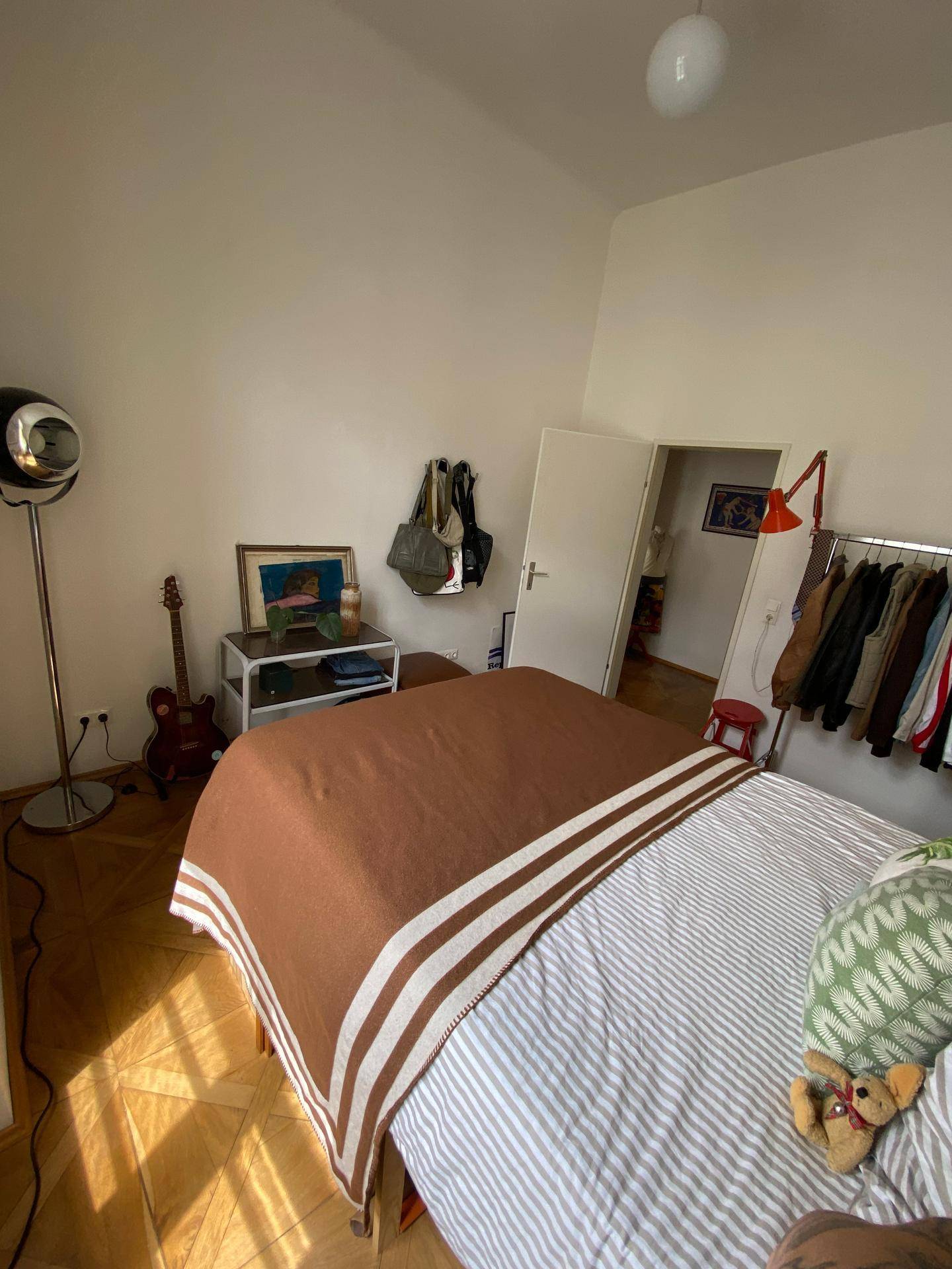 Schlafzimmer mit Holzboden, Kleiderstange und Zugang zu einem weiteren Raum.