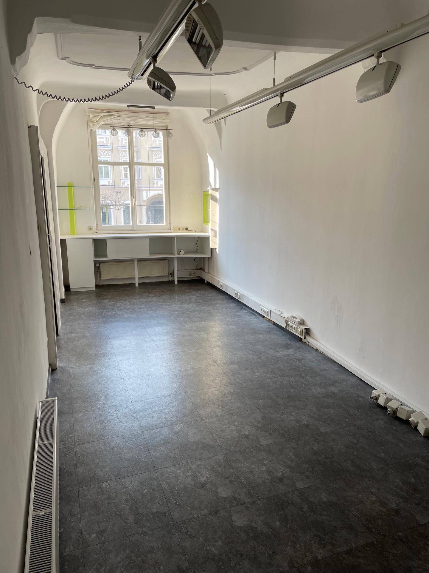Heller Innenraum mit dunklem Boden, weißen Wänden und einem Fenster, ausgestattet mit Deckenstrahlern und Heizkörper.